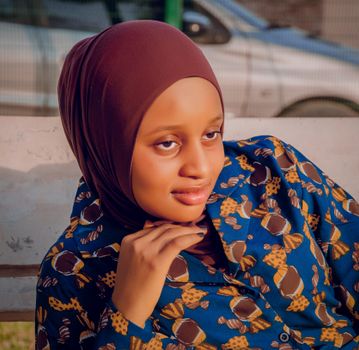 A young woman in a patterned blue shirt and hijab poses confidently outdoors.