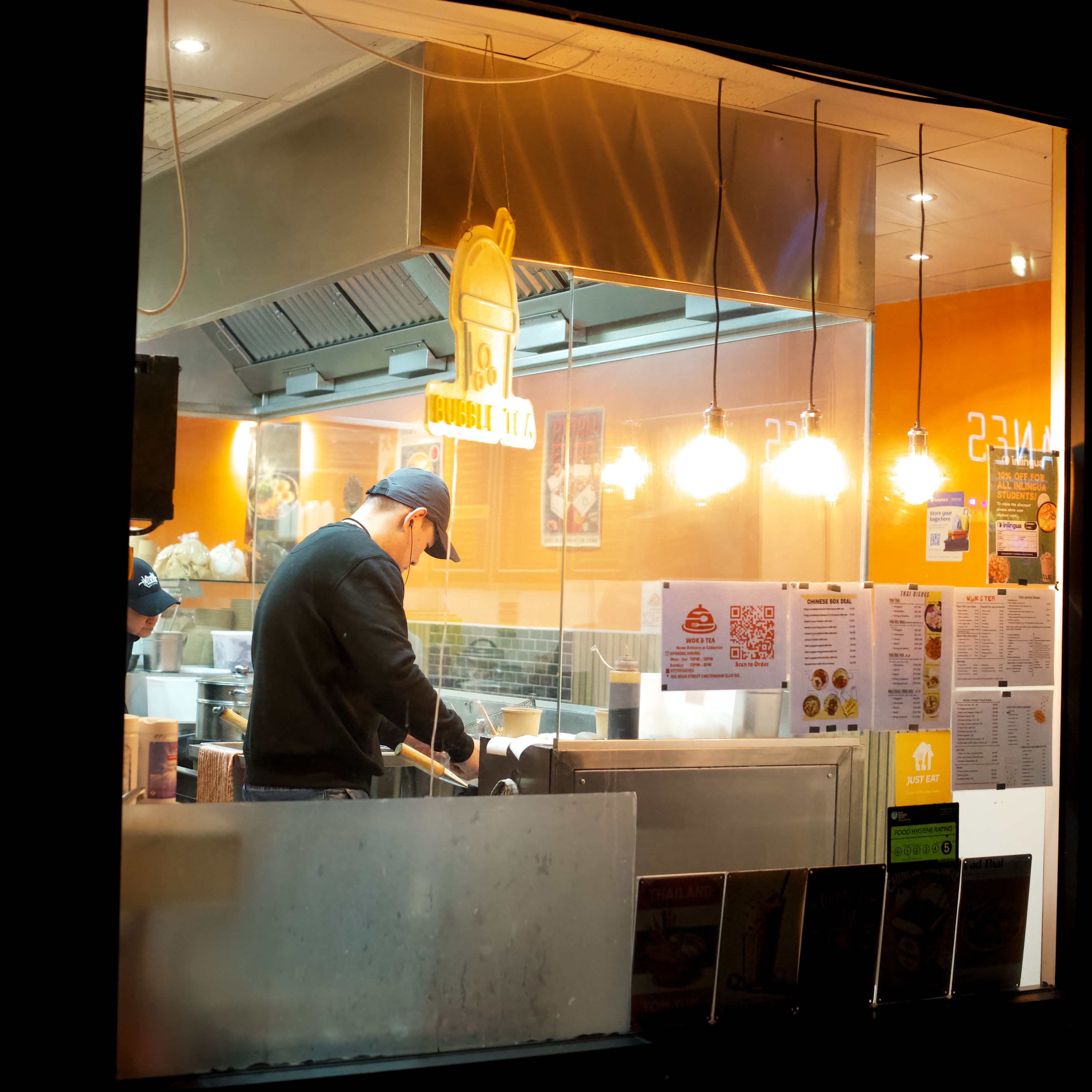 A chef works diligently behind a restaurant window, preparing food in a well-lit kitchen at night.