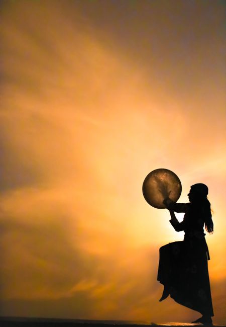 A silhouette of a woman playing a drum against a vibrant sunset sky.
