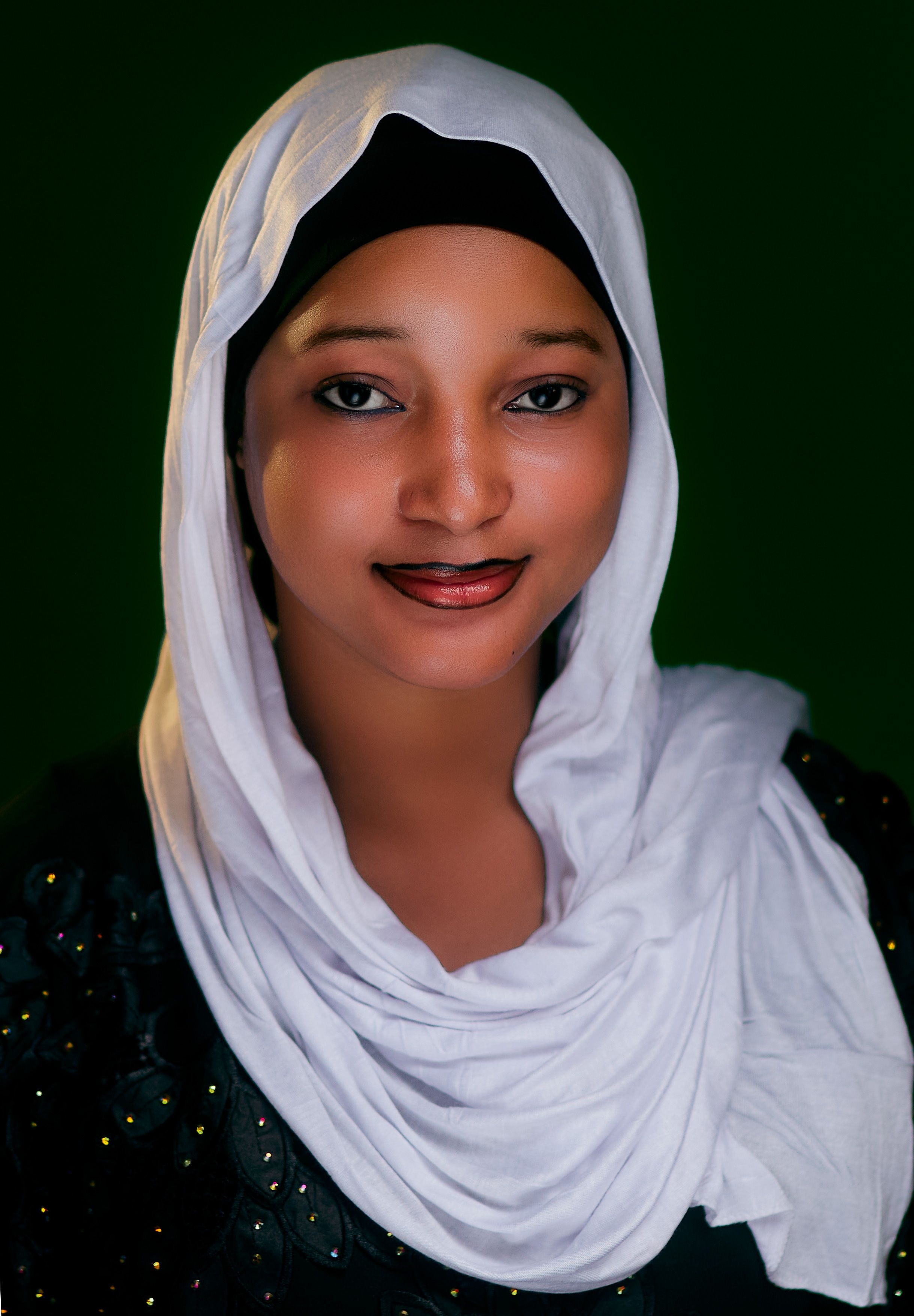 Portrait of a smiling woman in a white hijab with a dark green background.