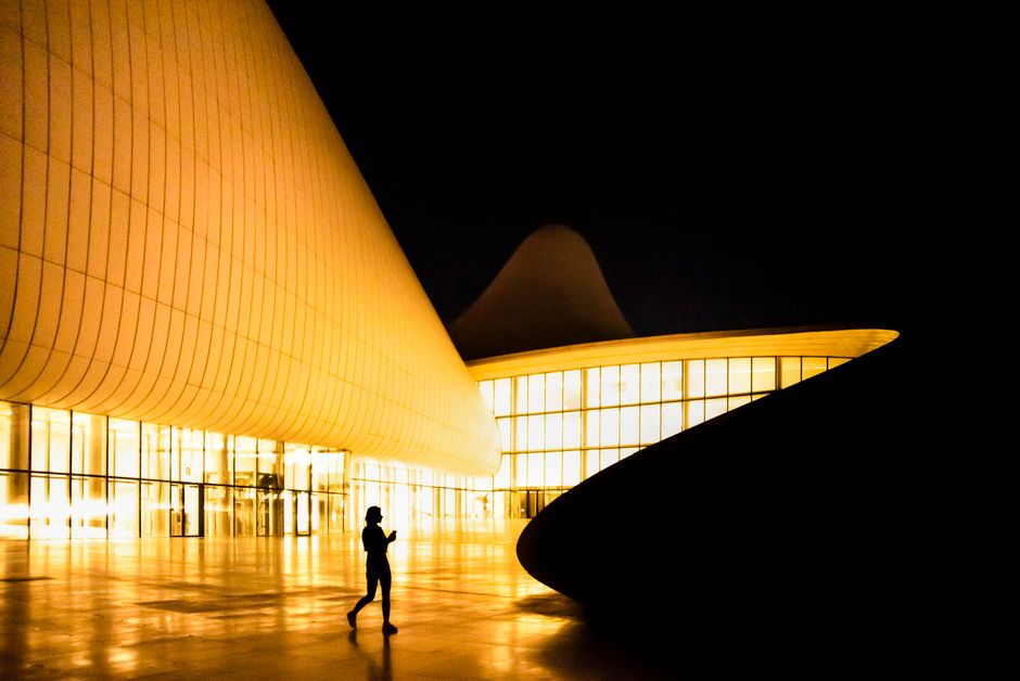 Silhouette of a person walking outside a brightly lit modern curved building at night.