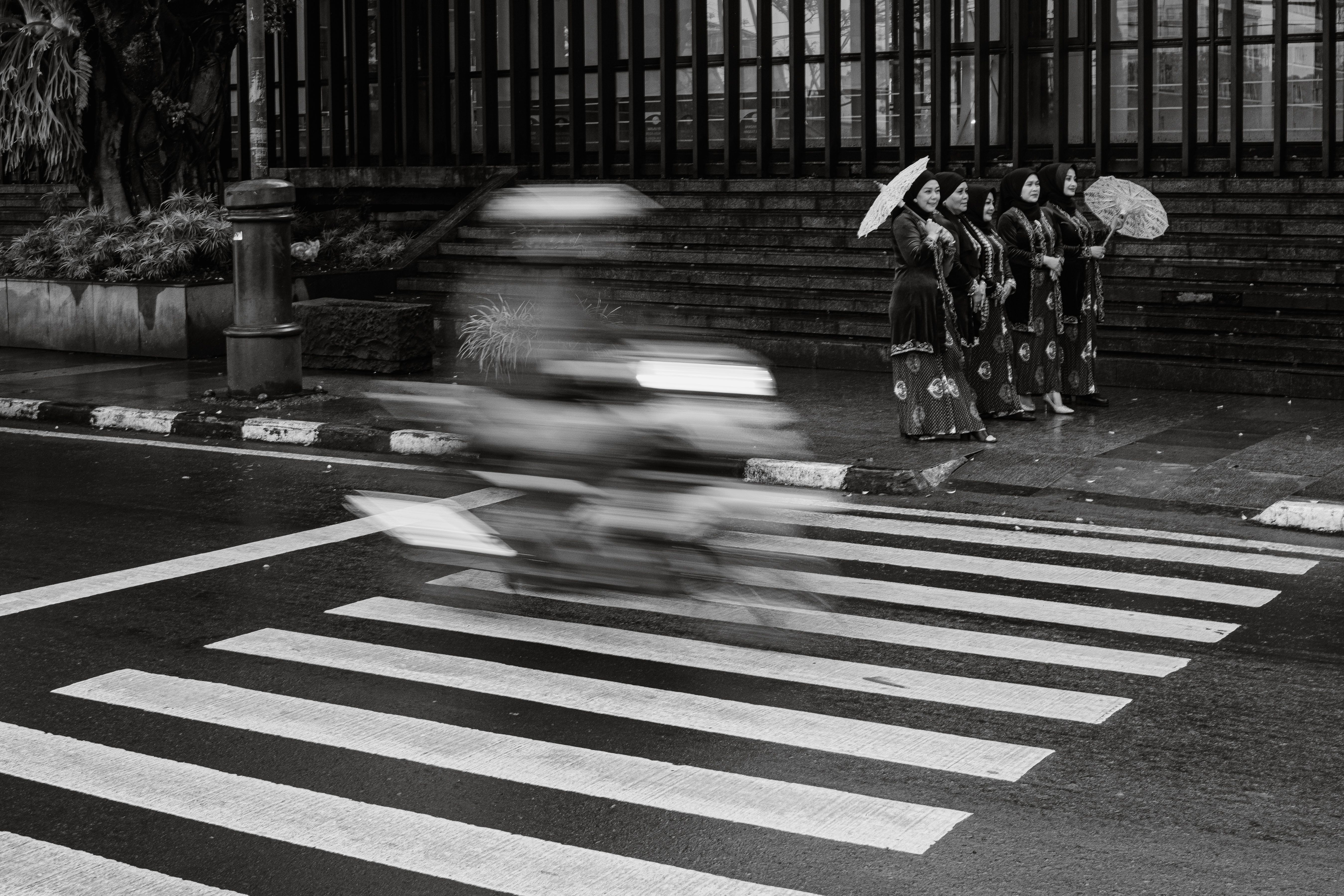 Black and white photo of women in traditional attire with umbrellas by a zebra crossing in Bandung, Indonesia.