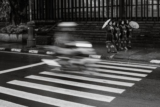 Black and white photo of women in traditional attire with umbrellas by a zebra crossing in Bandung, Indonesia.