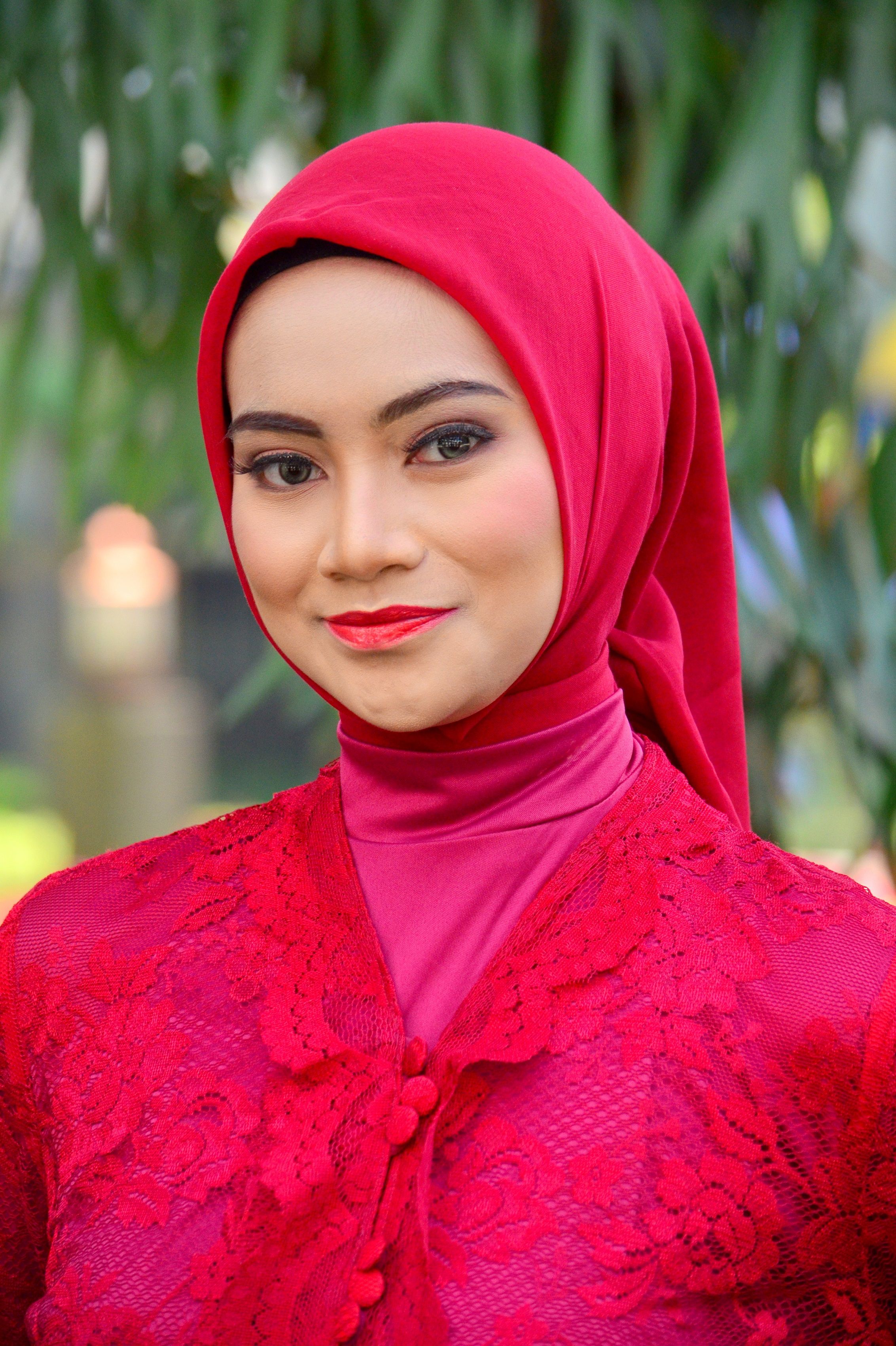 A woman dressed in an elegant red hijab and outfit smiles outdoors, capturing cultural beauty.