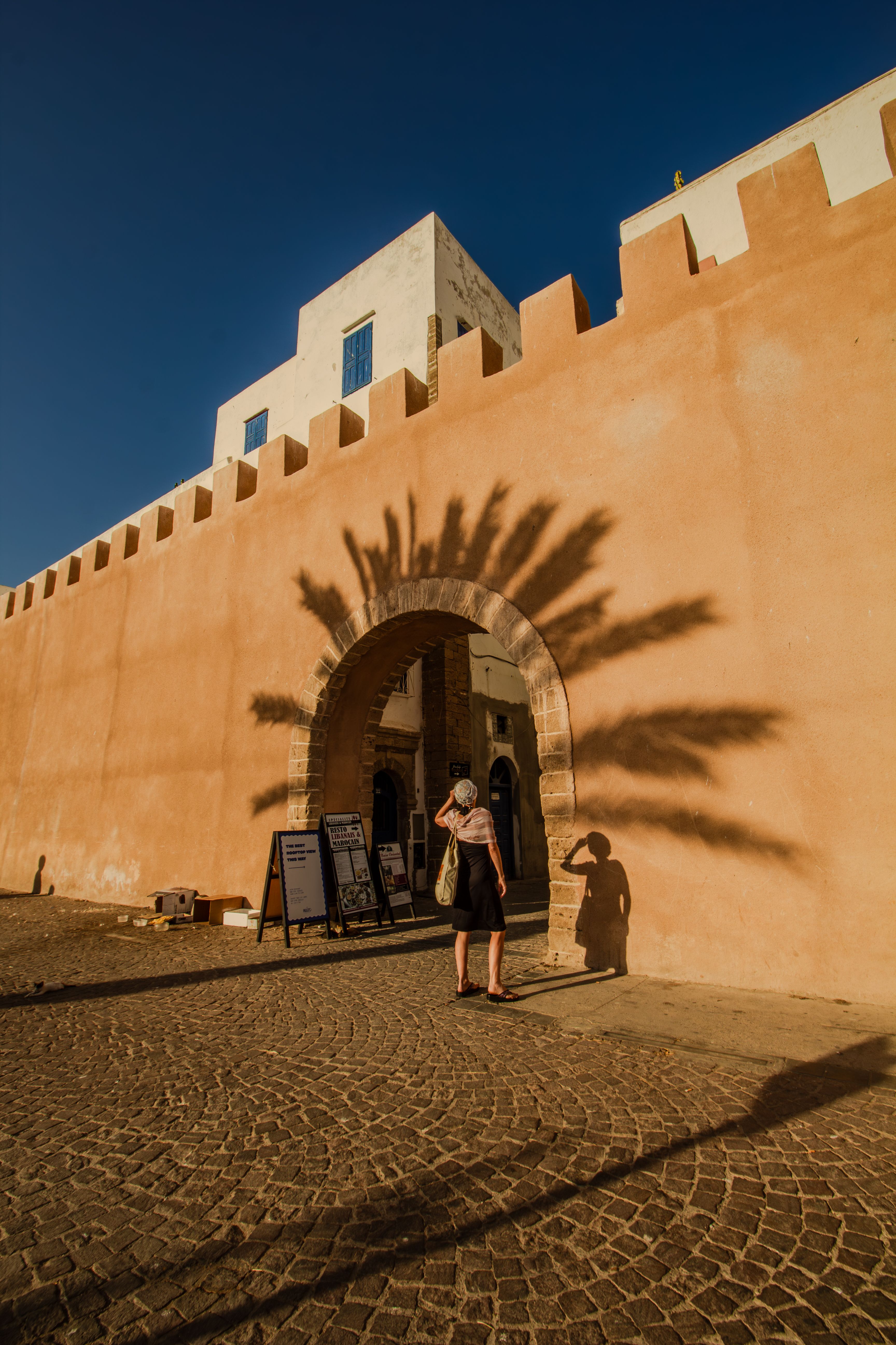 A person walks through a historic archway casting shadows in Essaouira, Morocco.