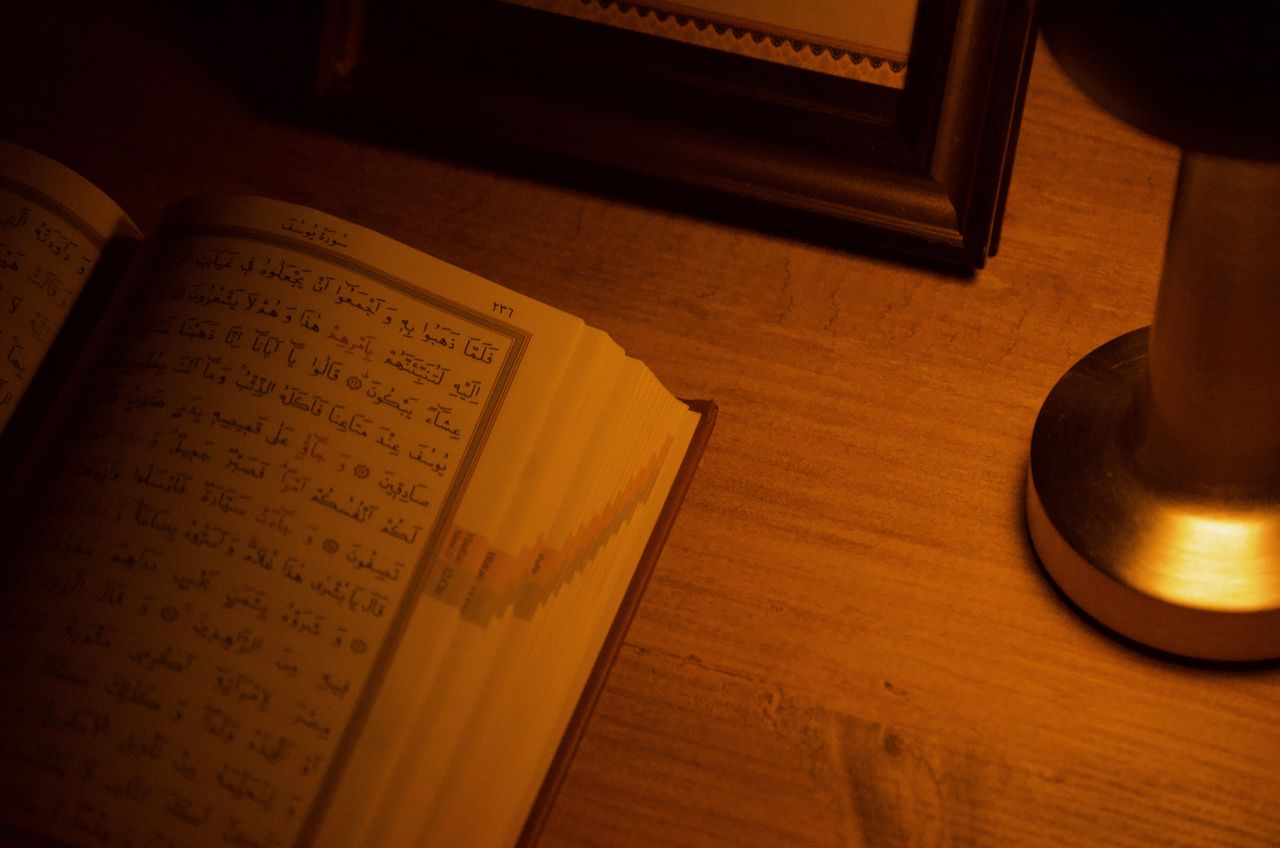 An open Quran on a wooden table next to a lit lamp, set in warm ambient light.