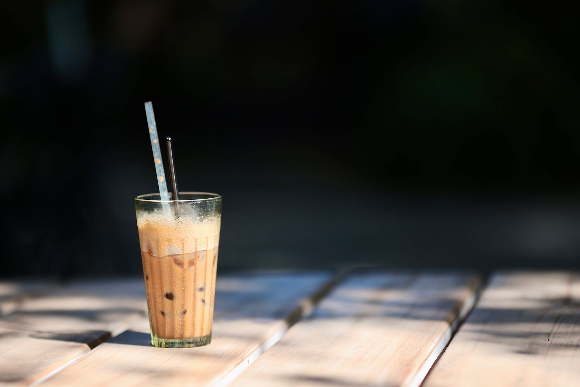 A glass of iced coffee with two straws sits on a wooden table in the sun.