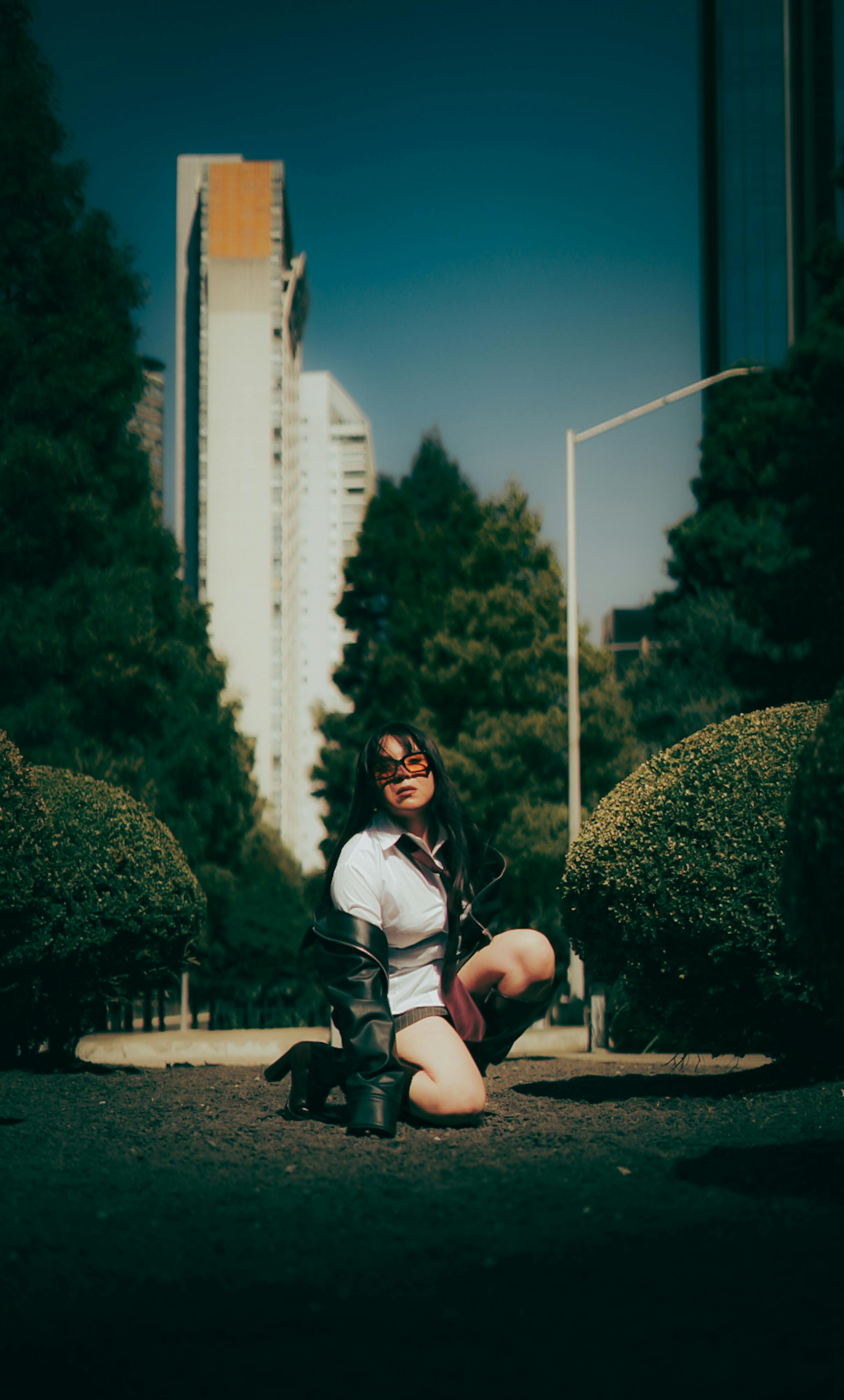 Ücretsiz Kadın, arka planda modern binaların bulunduğu bir şehir parkında diz çökmüş halde, doğa ve kentsel yaşam arasında bir tezat oluşturuyor. Stok Fotoğraflar