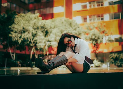 Trendy woman in boots and sunglasses sits by a modern building in a relaxed pose. Urban fashion scene.