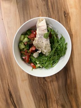 Fresh salad bowl with arugula, tomatoes, cucumbers, and grilled chicken on a wooden table.