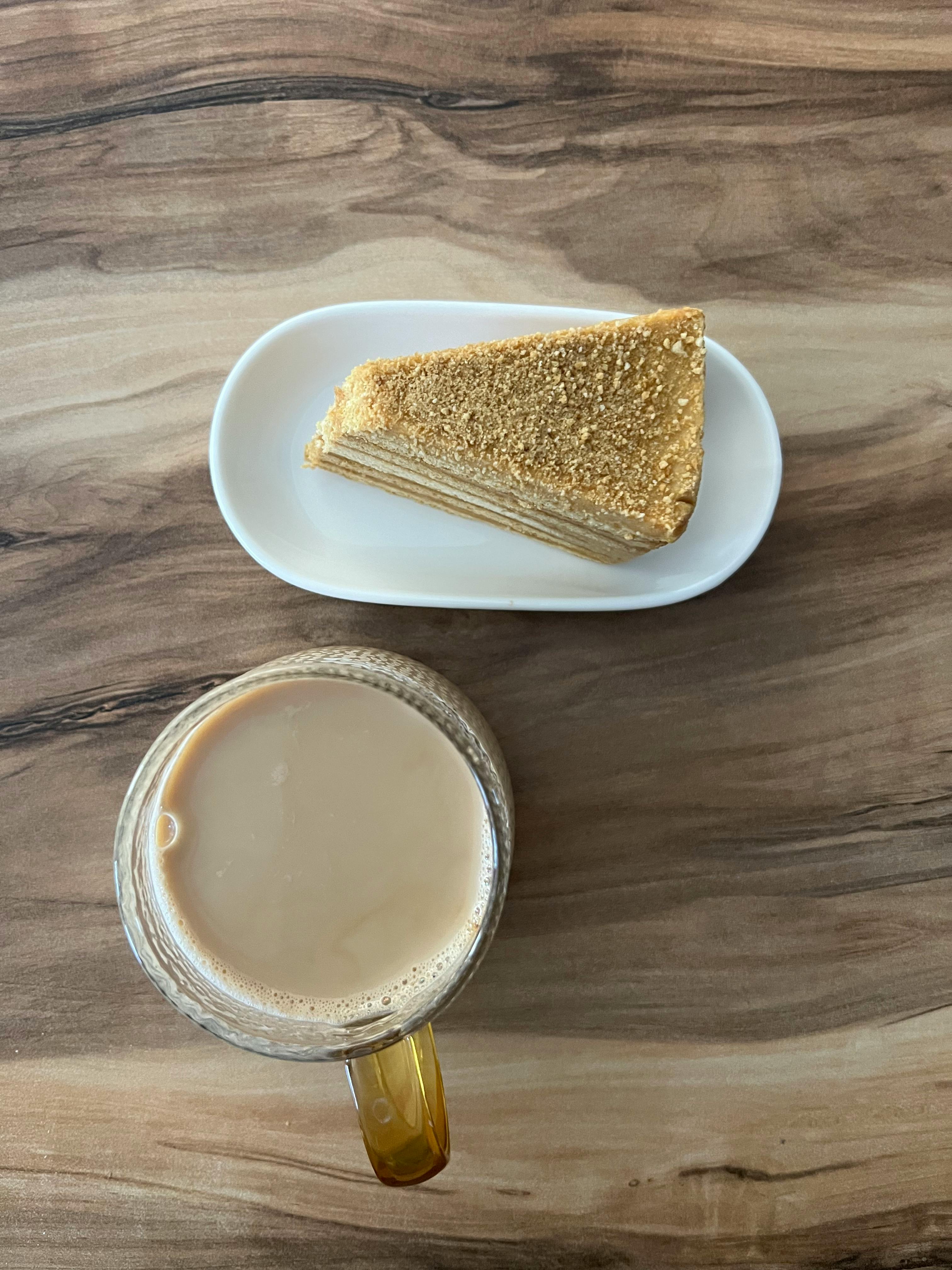 A slice of layered cake on a white plate with a glass of coffee on a wooden table.