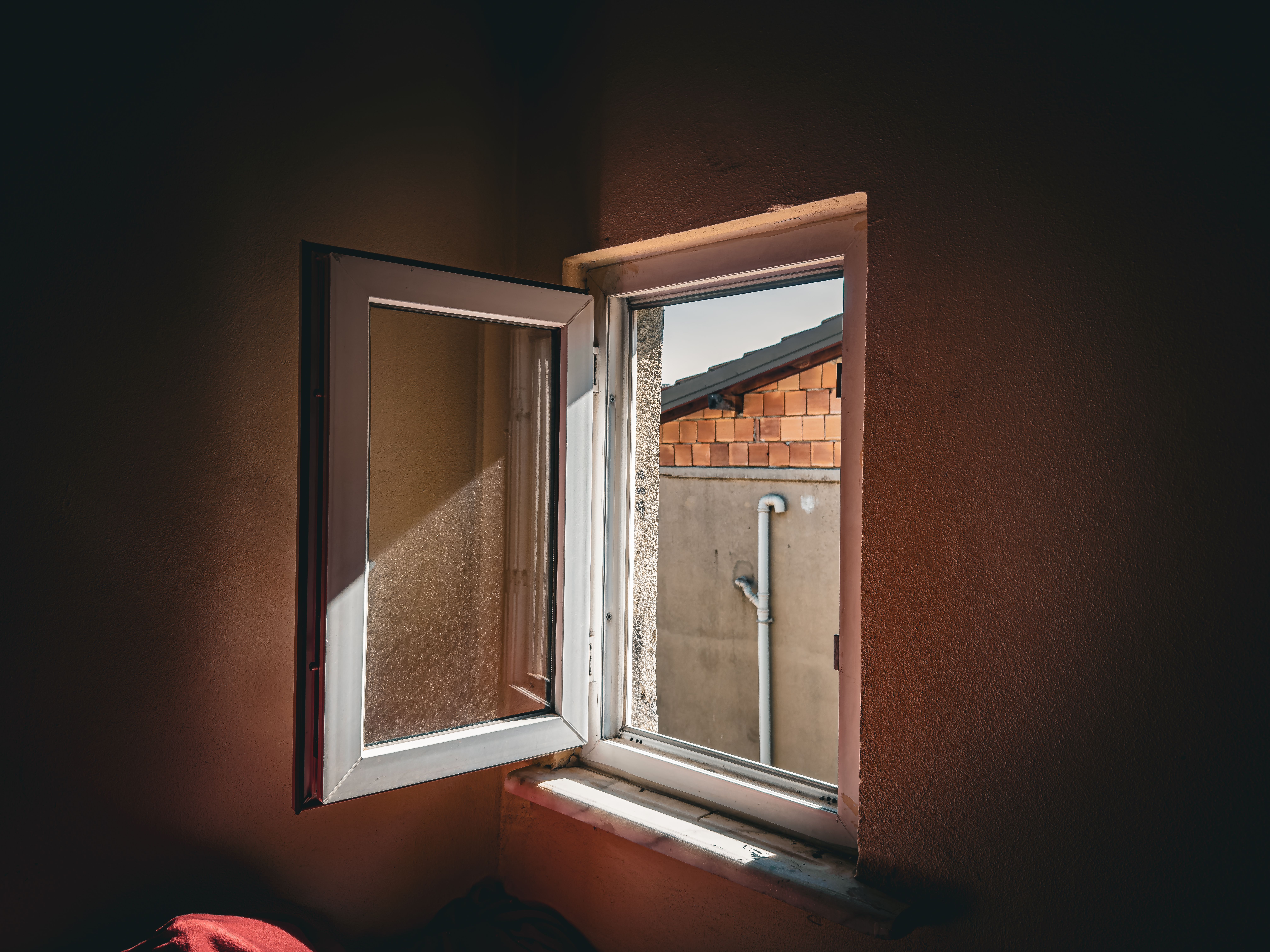 Open window view of a brick wall and pipe, sunlight casting shadows indoors.