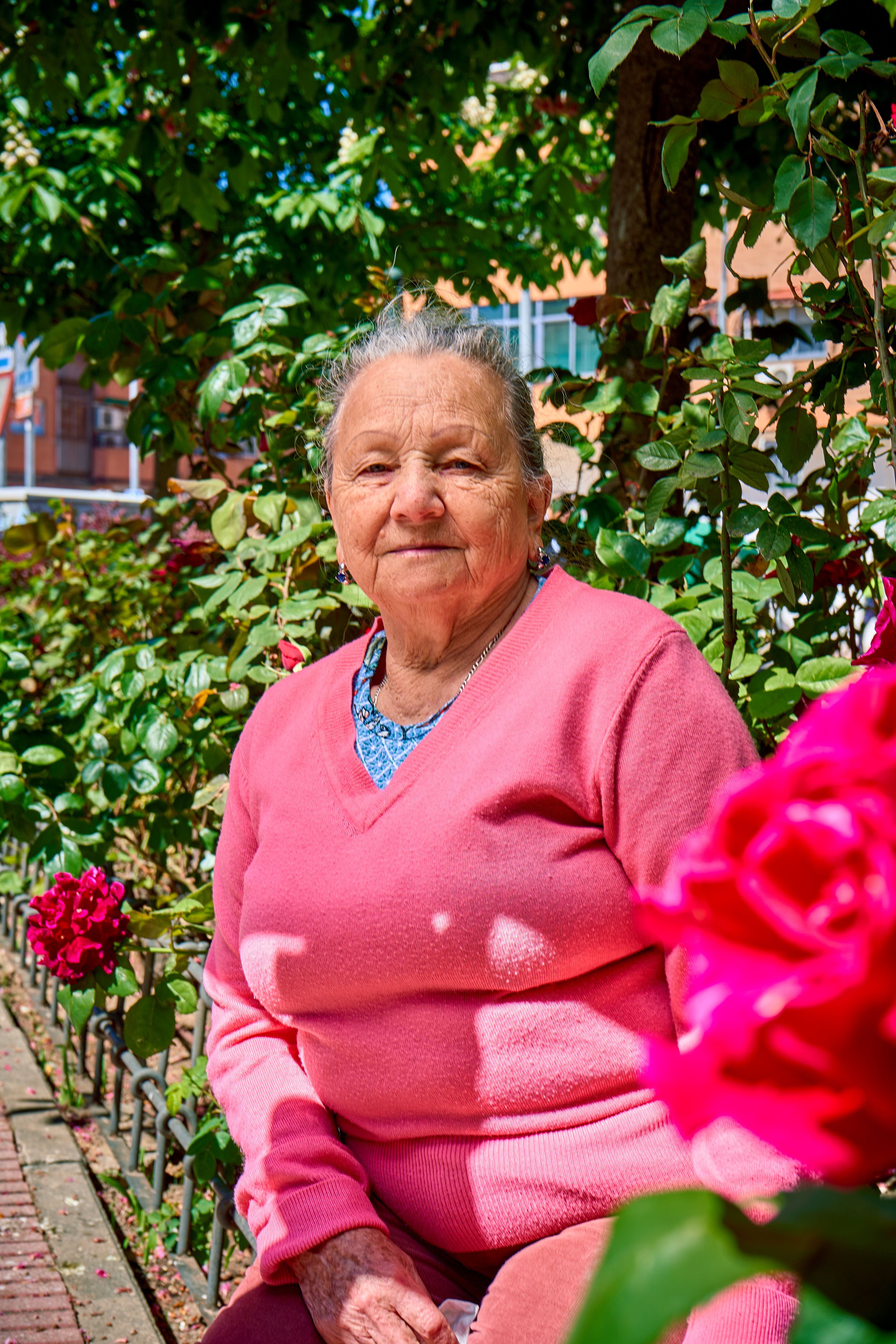 Elderly woman in a garden setting with blooming flowers on a sunny day.