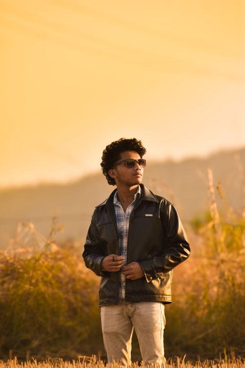 Free Man in a leather jacket standing in a field during golden hour, exuding confidence. Stock Photo