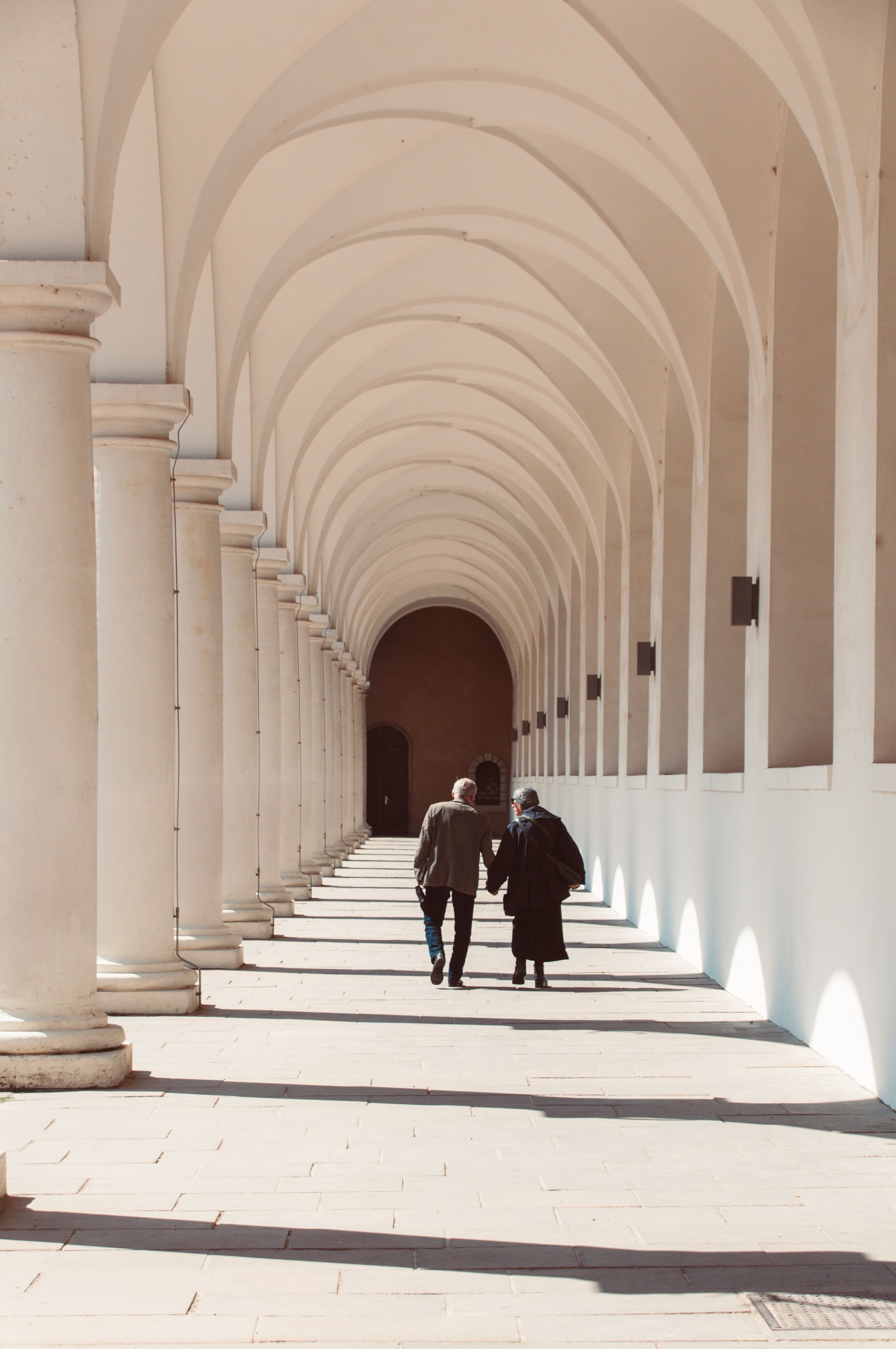 An elderly couple strolls down a sunlit, arched corridor, symbolizing love and serenity.