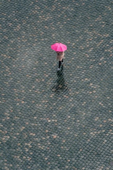 A person stands with a pink umbrella on a wet cobblestone path, captured from above.