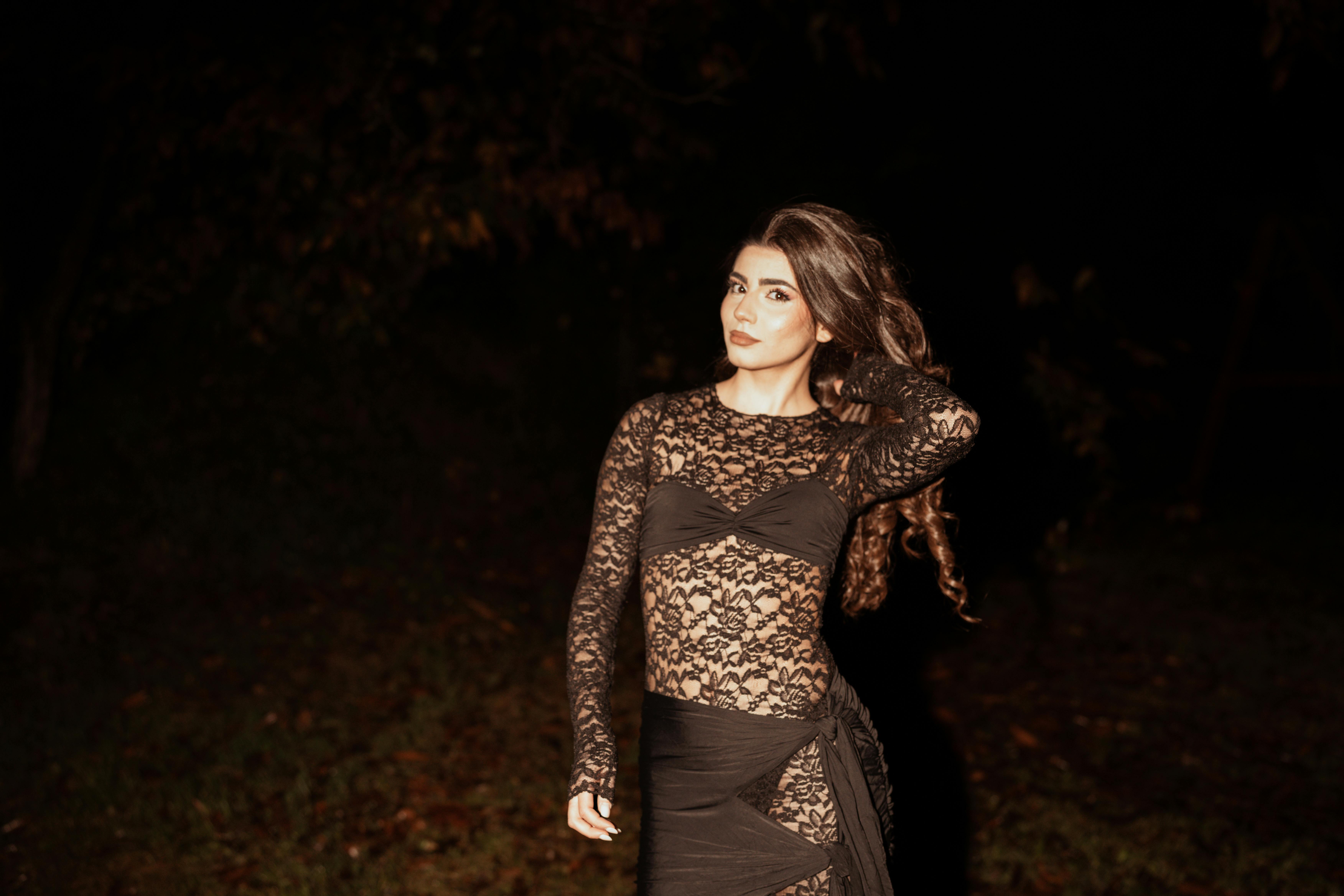 A woman poses elegantly in a black lace dress during nighttime, showcasing fashion and style.