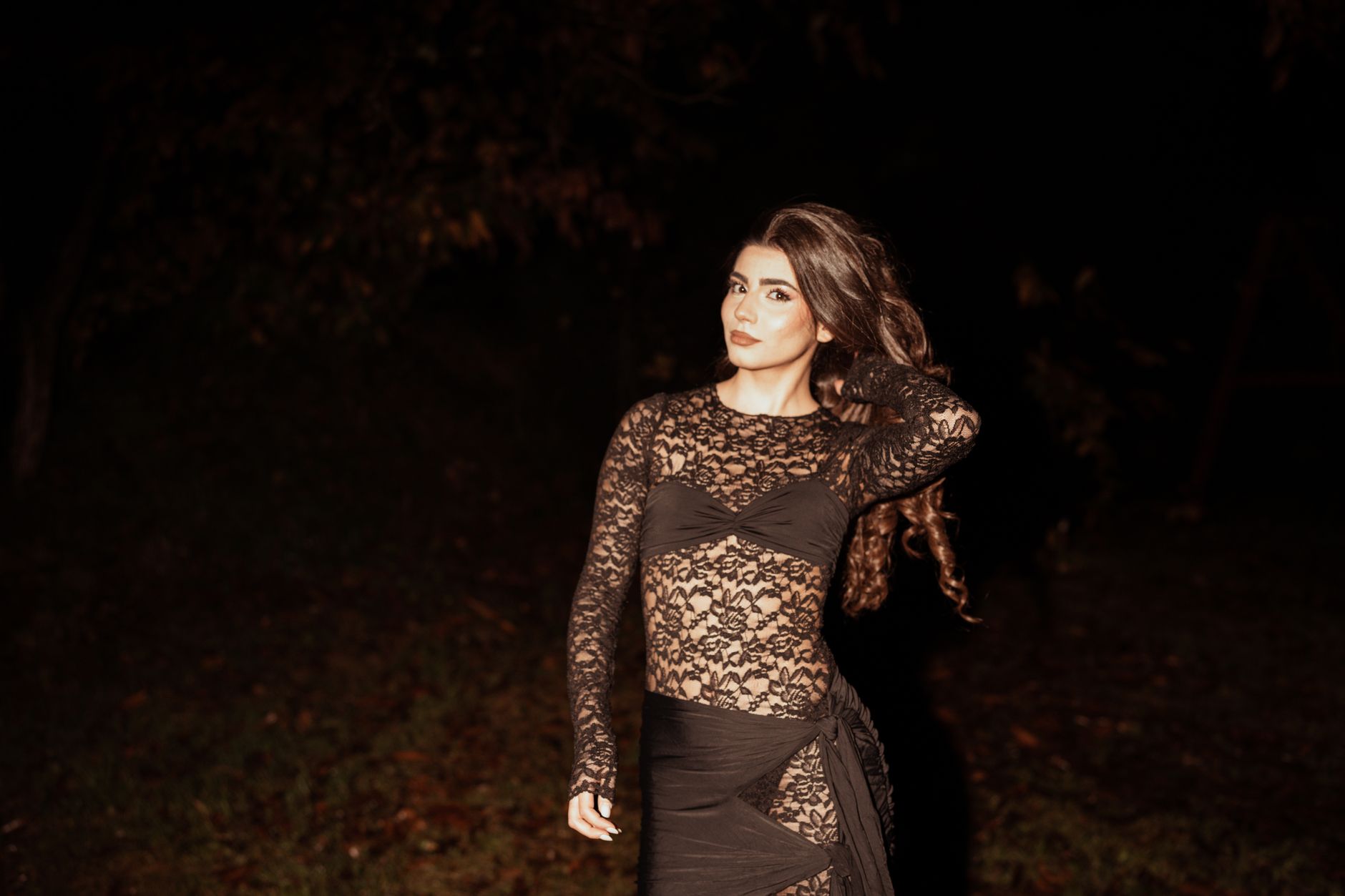 A woman poses elegantly in a black lace dress during nighttime, showcasing fashion and style.