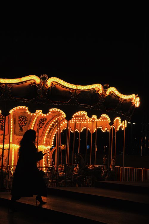 Free A silhouette of a woman walking past a brightly lit carousel during the night, creating a contrasting visual. Stock Photo