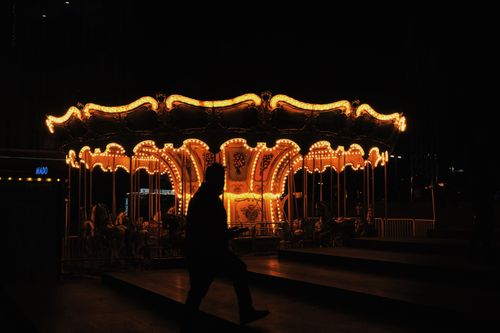 Free A person walks past a brightly lit carousel, creating a striking silhouette at nighttime. Stock Photo
