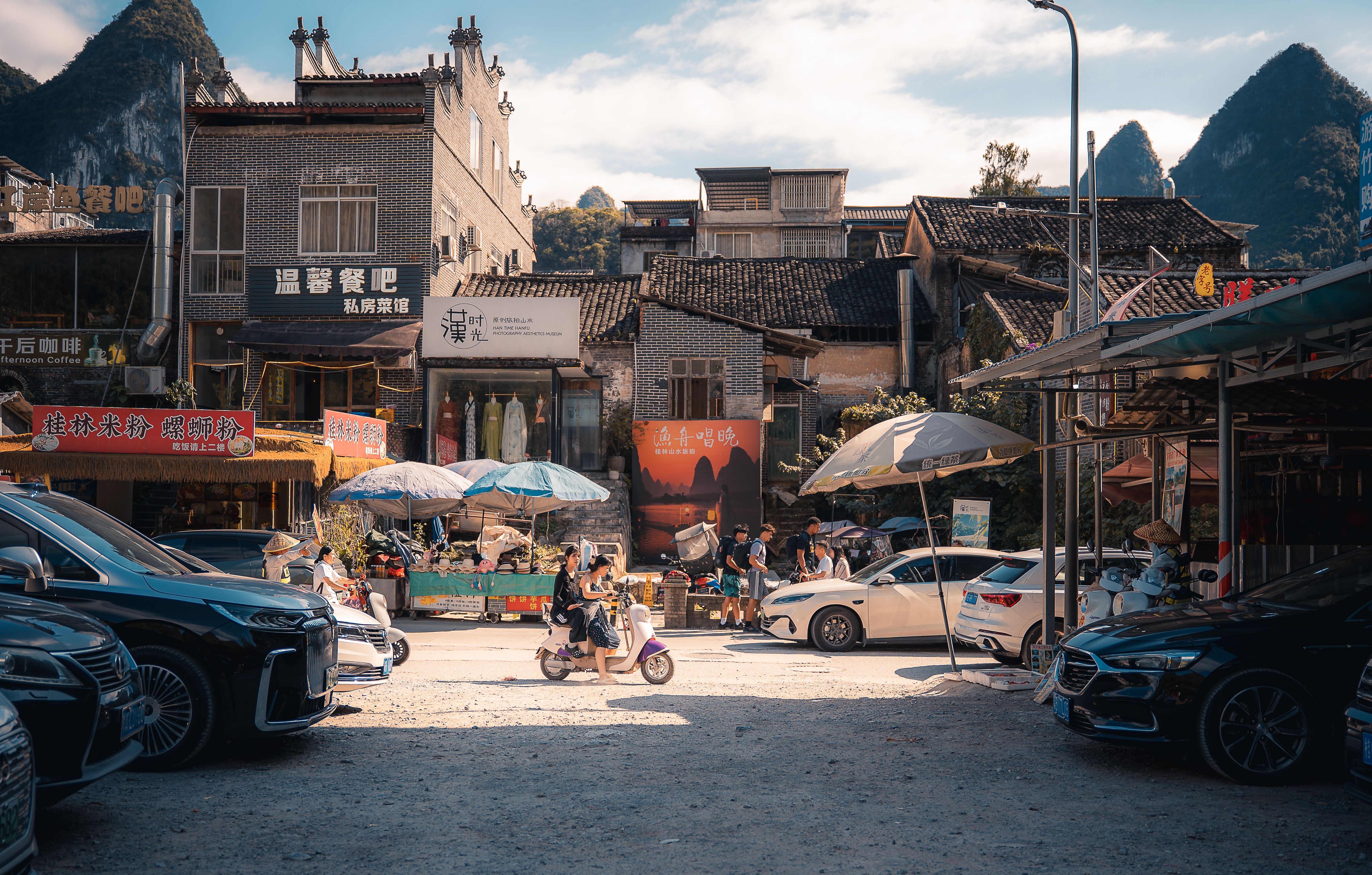 Street scene in Xingping, China featuring parked cars, motorcycles, and local shops under a sunny sky