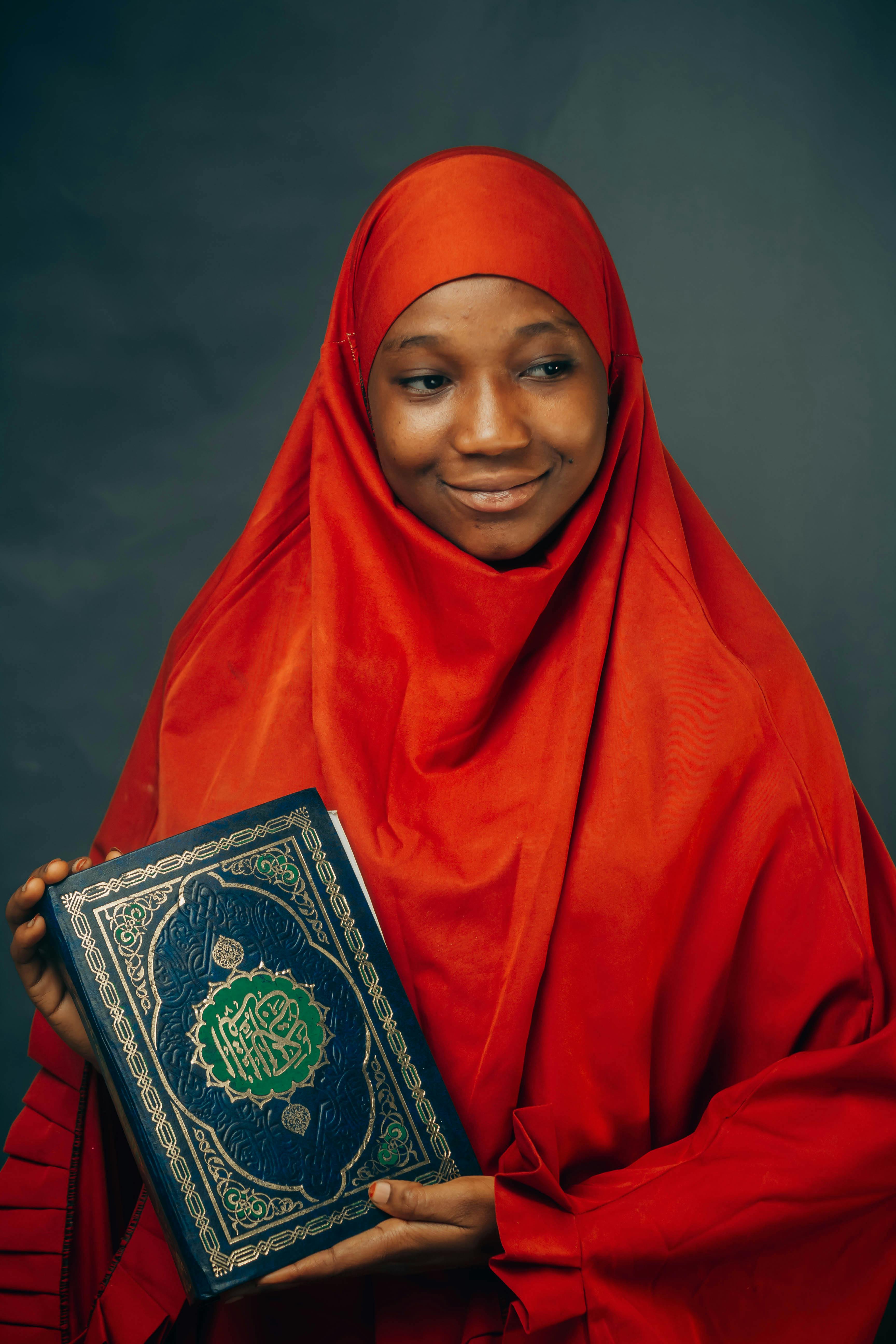 Portrait of a young woman in a red hijab holding a Quran, smiling softly.