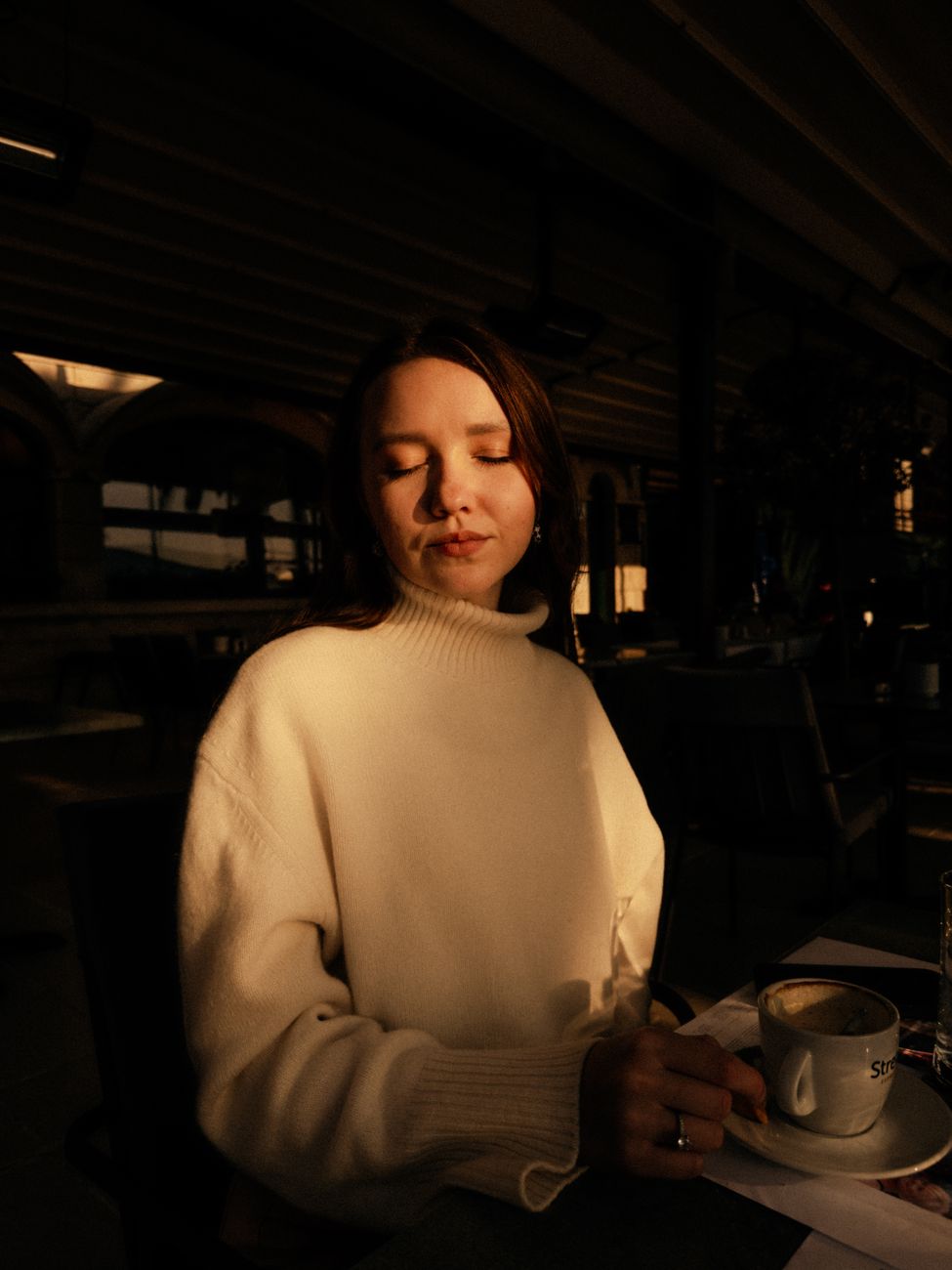 A woman basks in warm sunset light while enjoying coffee indoors, creating a peaceful ambiance.