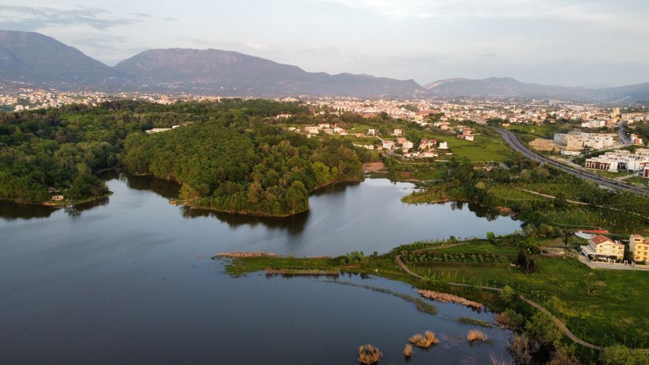 Scenic aerial photo of Tirana, Albania with lush greenery, lake, and distant mountains at daytime.