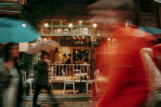 Blurred pedestrians with umbrellas pass a charming café with warm ambiance and vintage decor.
