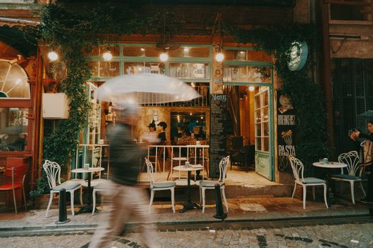 Cozy street cafe with vintage decor, blurred pedestrian passing by with umbrella, urban vibes.