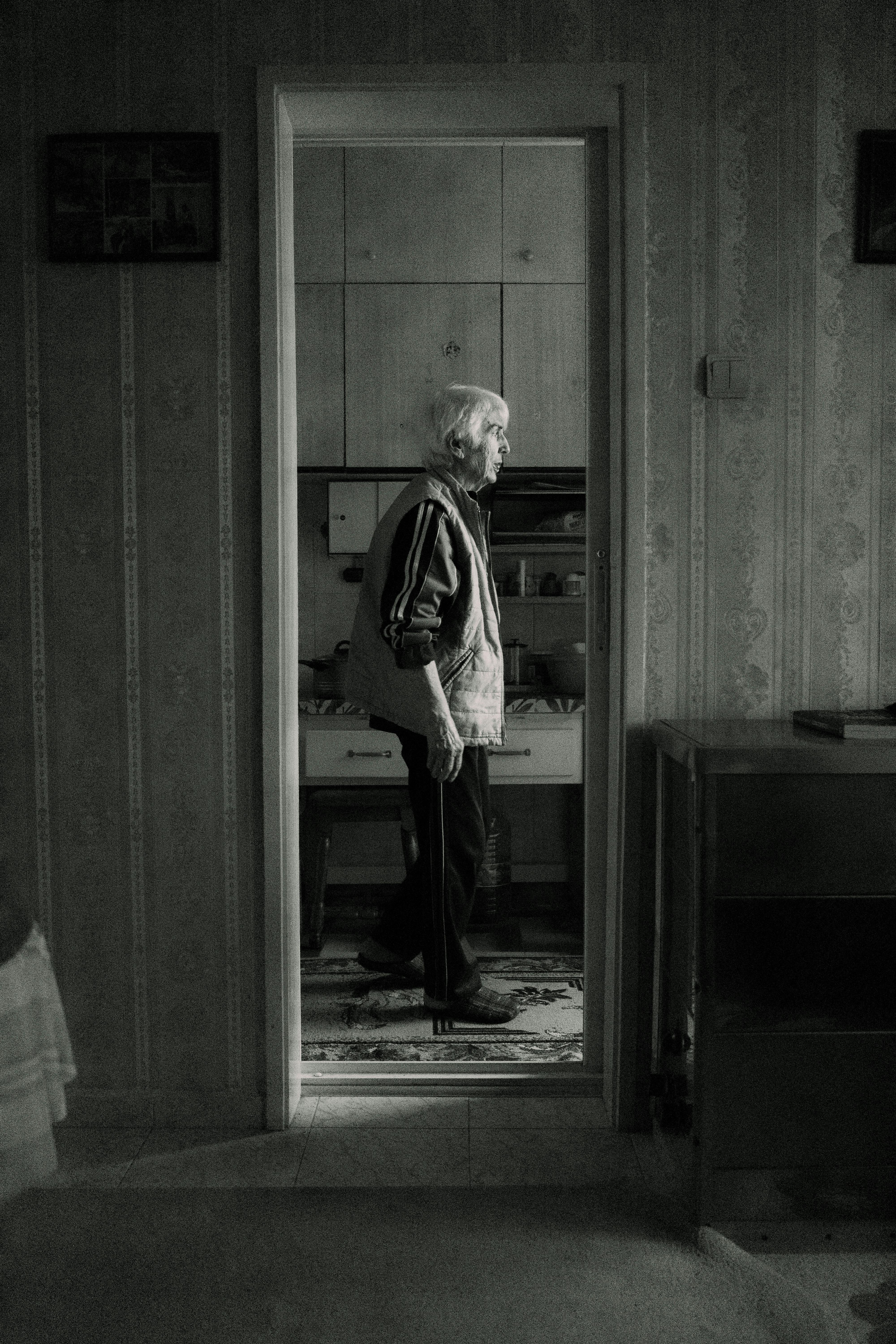 Free Black and white photo of an elderly woman standing in an interior doorway, conveying solitude. Stock Photo