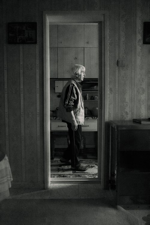 Free Black and white photo of an elderly woman standing in an interior doorway, conveying solitude. Stock Photo
