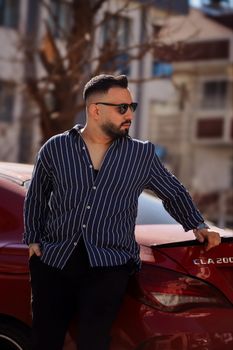 Man in striped shirt leans on red car in city street, exuding urban style and confidence.