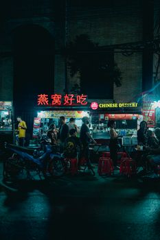 A bustling street market at night with a vibrant Chinese dessert stall and people enjoying food.