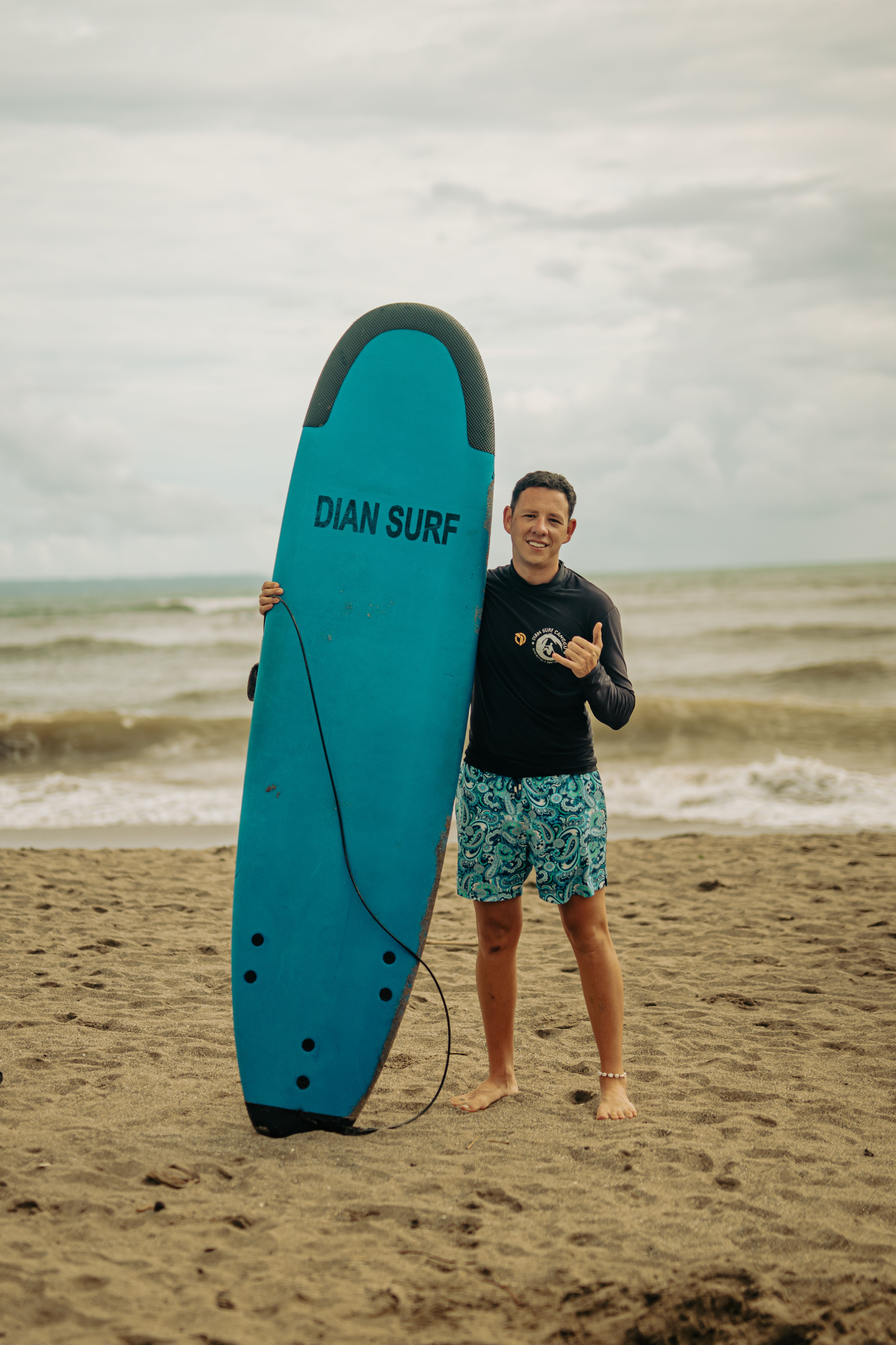 Surfer posing with a board on a sandy beach, cloudy day, relaxed vibe.