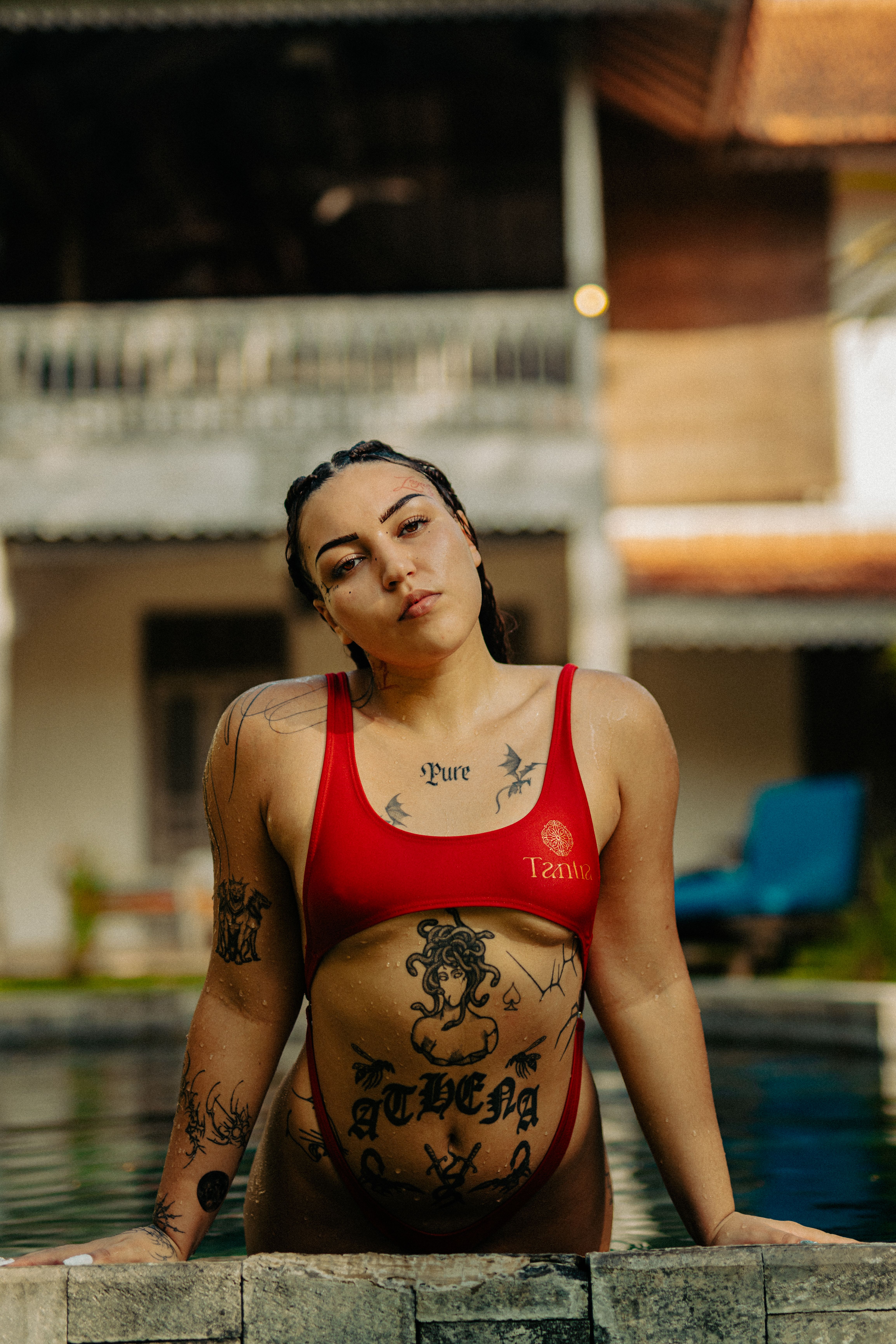 Woman in a red bikini leaning on the poolside exuding confidence, with visible intricate tattoos, outdoors.