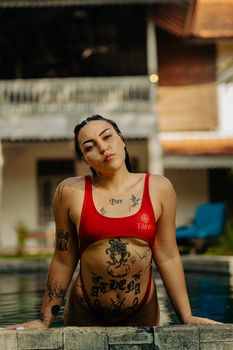 Woman in a red bikini leaning on the poolside exuding confidence, with visible intricate tattoos, outdoors.