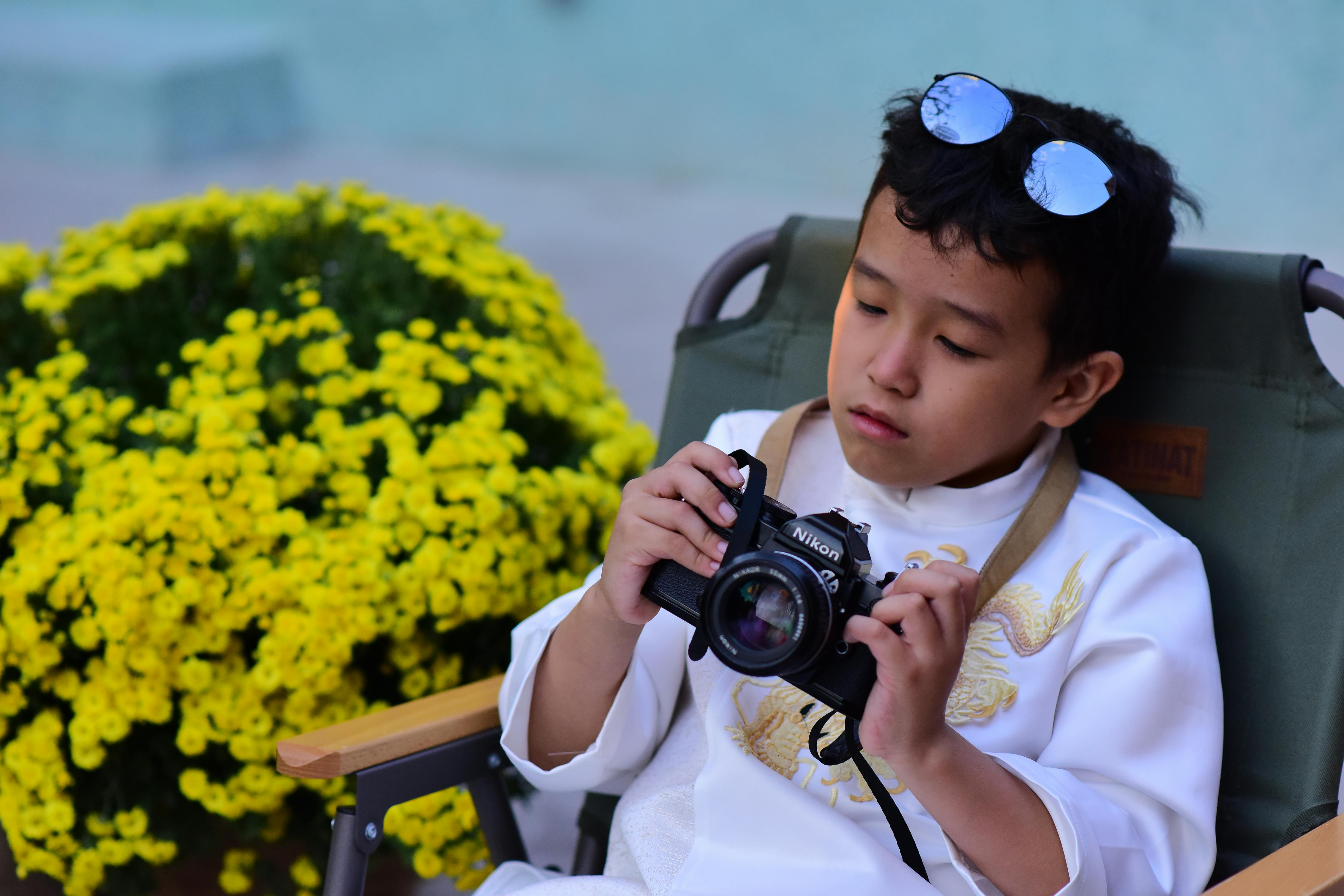 A young boy in traditional clothing examines a vintage camera beside yellow flowers outdoors.