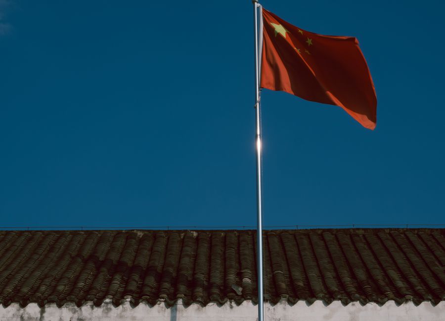 Chinese flag waving over traditional building roof against a clear blue sky.