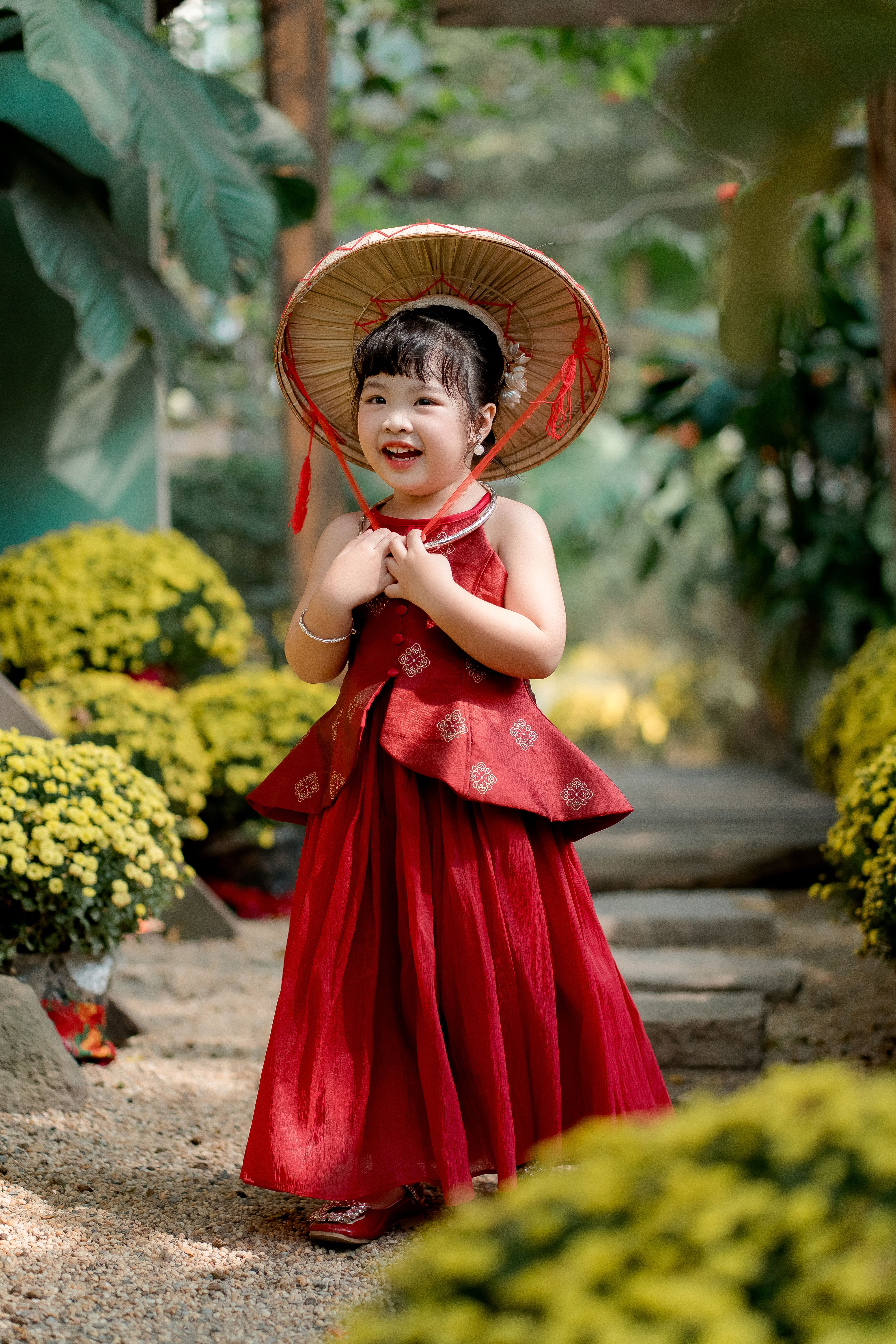 Smiling child in a red traditional dress with a bamboo hat in a garden setting.