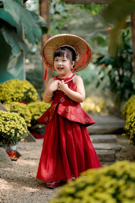 Smiling child in a red traditional dress with a bamboo hat in a garden setting.