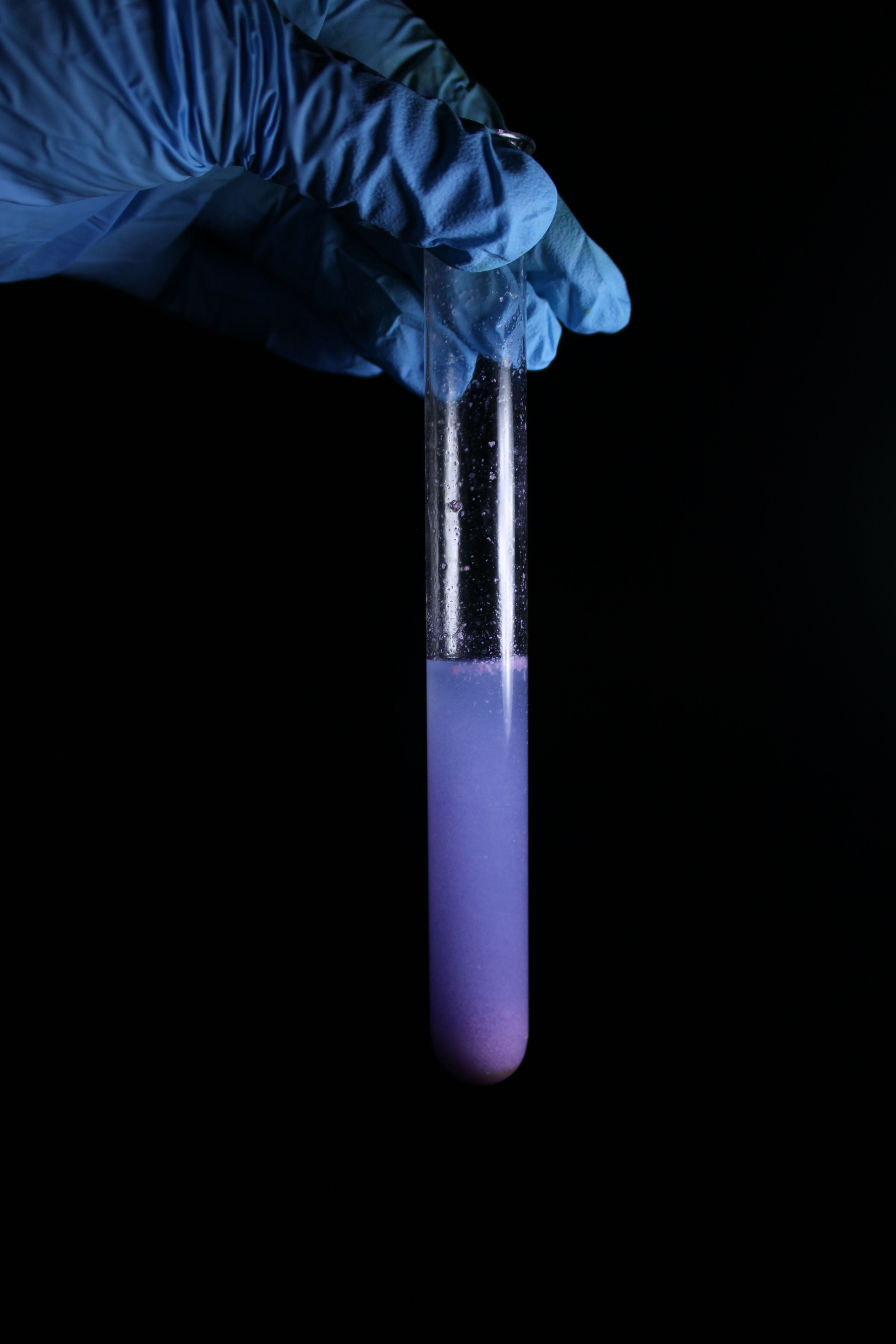 Close-up of gloved hand holding test tube with purple liquid against a black background.