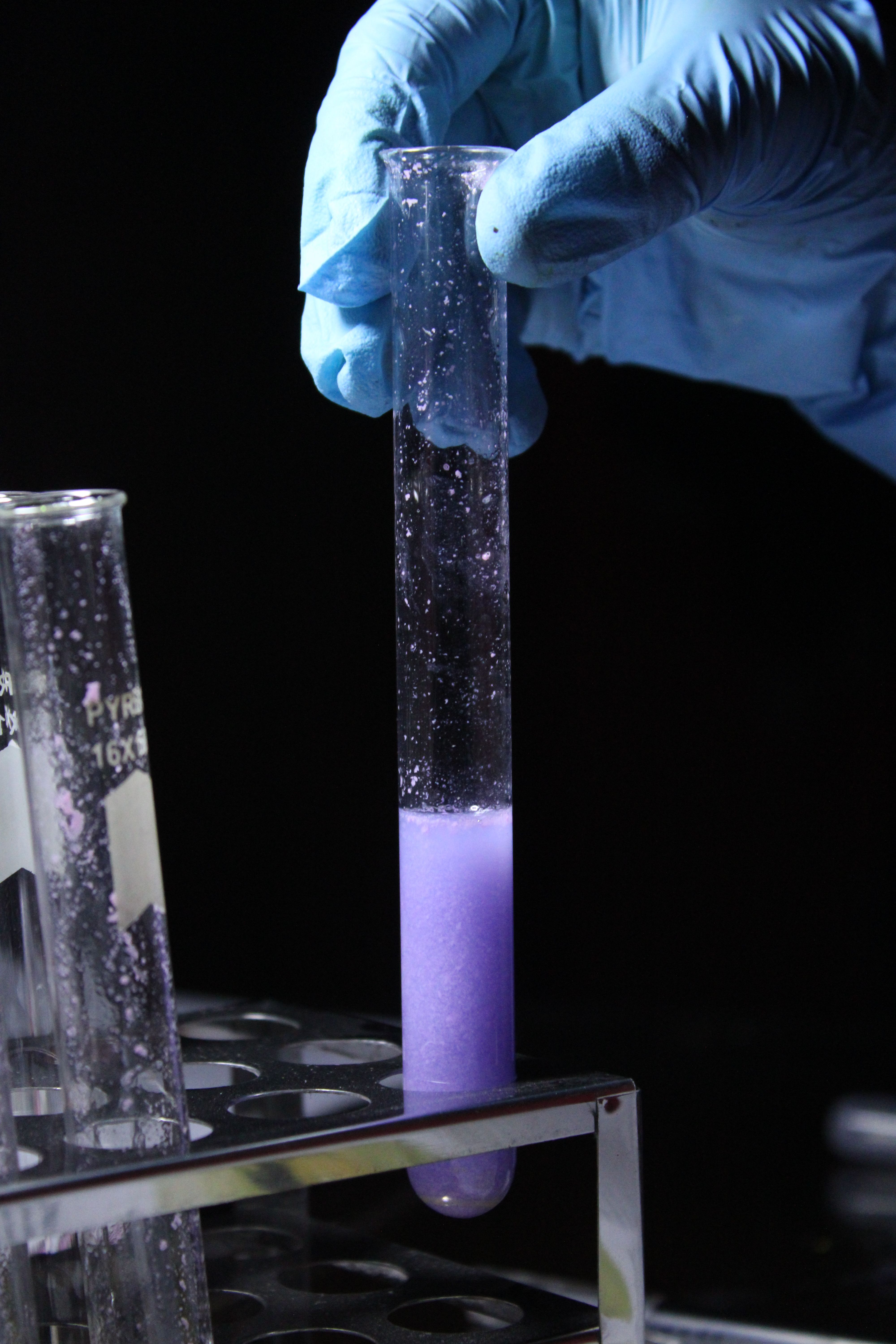 A scientist's gloved hand holding a test tube with purple liquid in a lab setting.