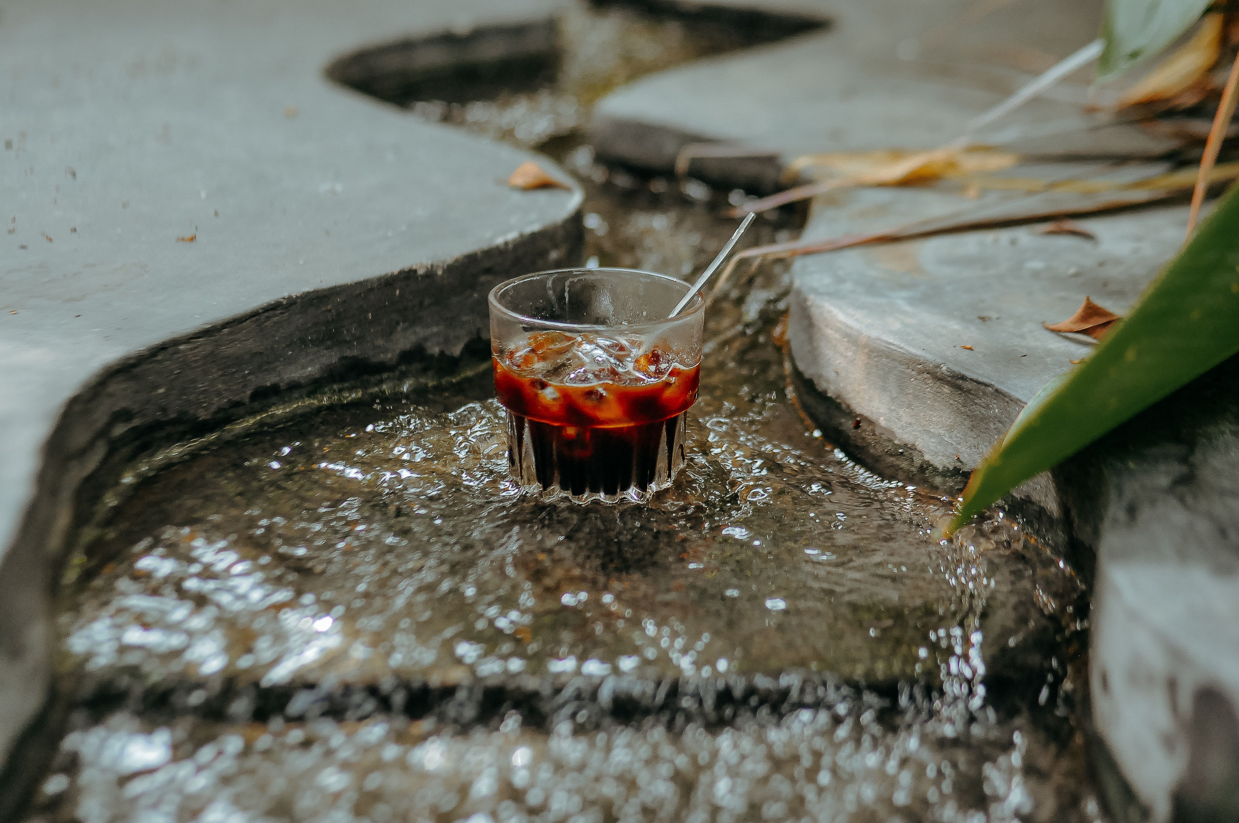 A glass of iced coffee sits in a small stream surrounded by natural stone outdoors.