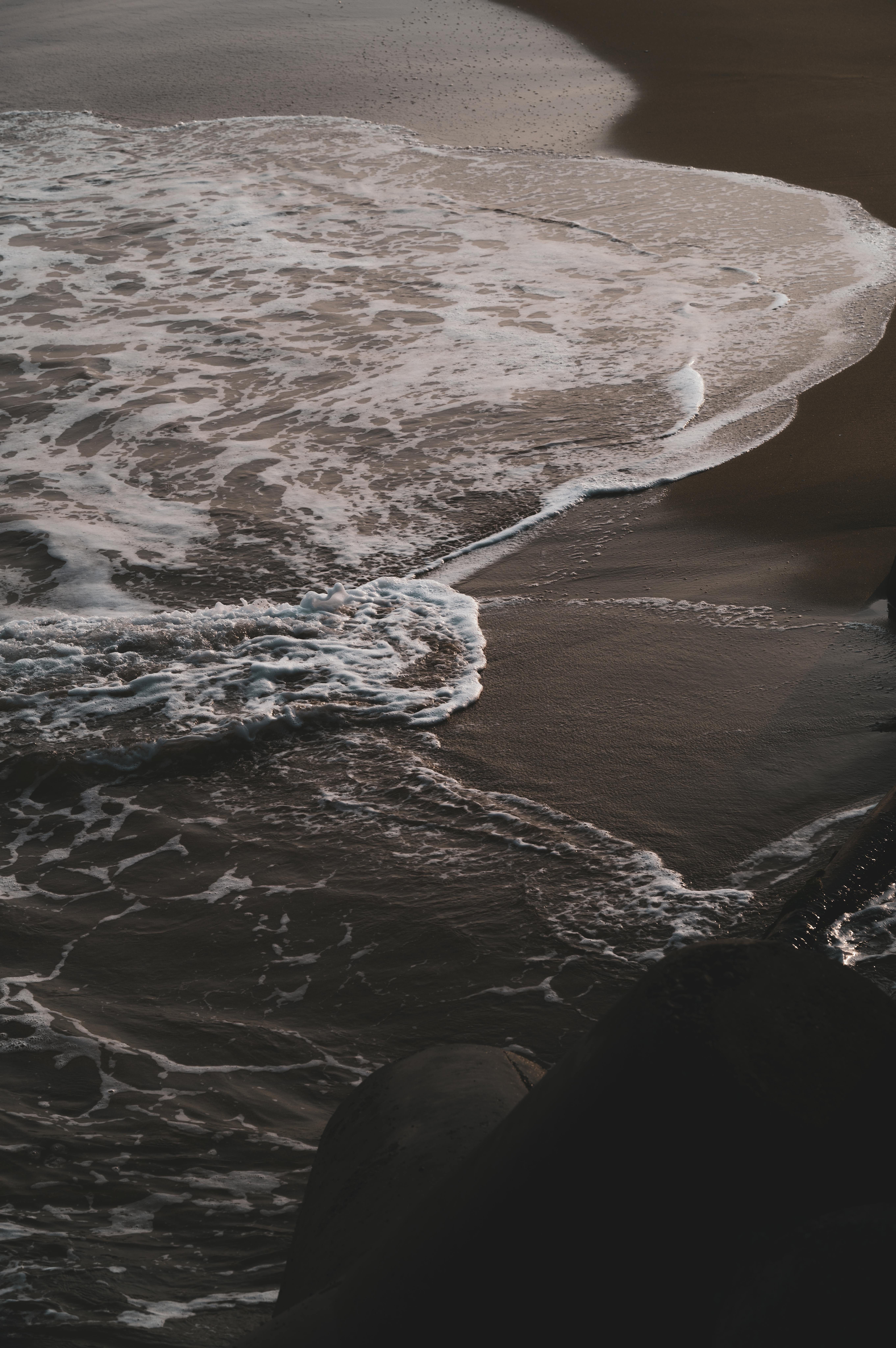 Ücretsiz Gün batımında kumlu sahile çarpan karanlık ve kasvetli okyanus dalgaları, sanatsal desenler oluşturuyor. Stok Fotoğraflar