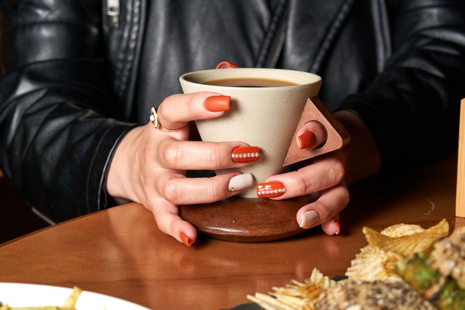 Close-up of manicured hands holding a coffee cup on a wooden table with snacks.