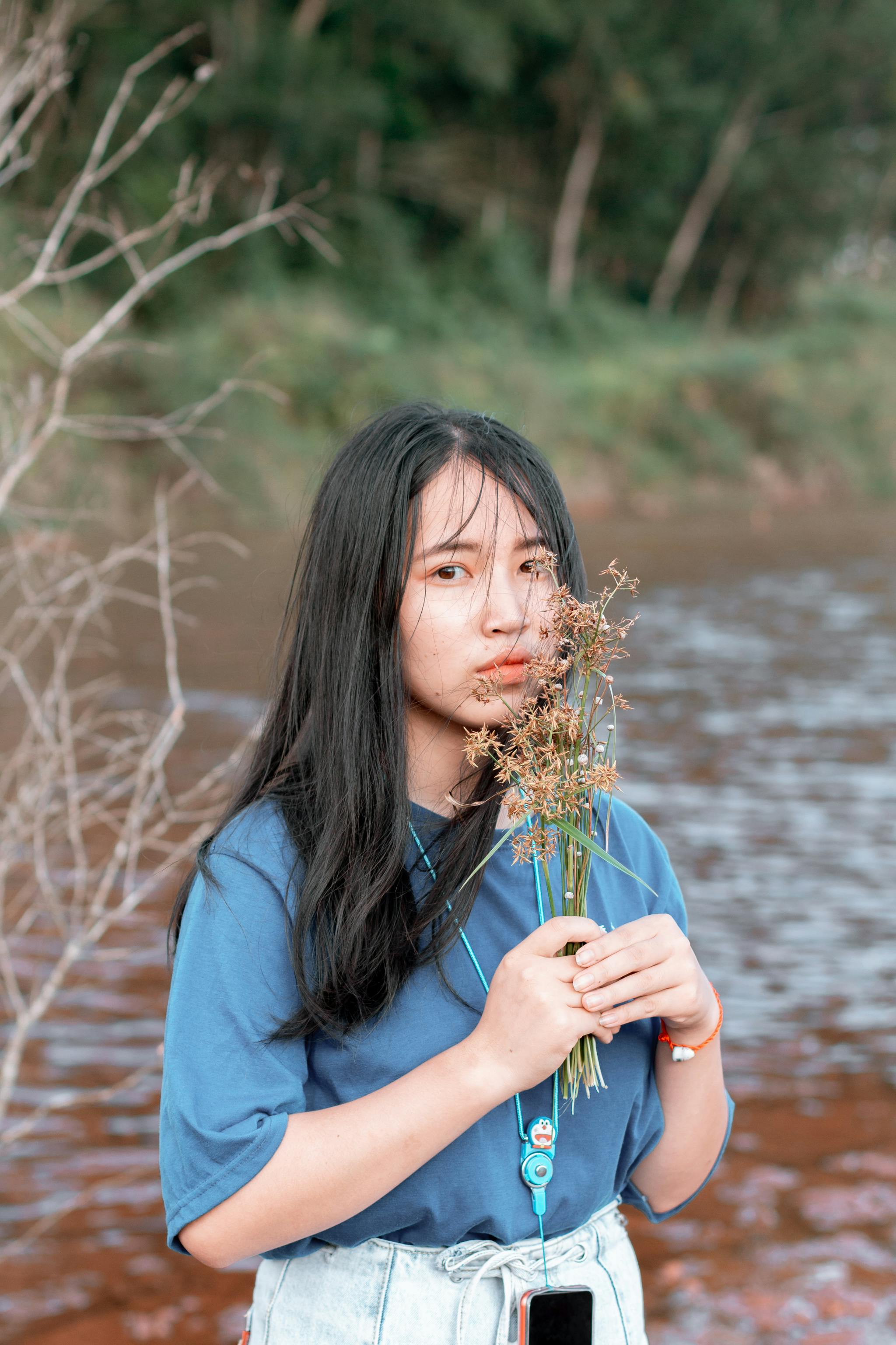 白天，一位身穿蓝色衬衫的年轻女子在宁静的河岸边捧着野花