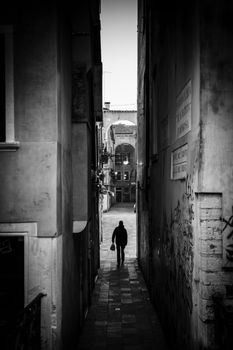 Black and white photo of a person walking in a narrow Venetian alley, creating a dramatic and moody atmosphere.