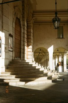 Sunlit historic arcade with stone steps and arches in a European city.
