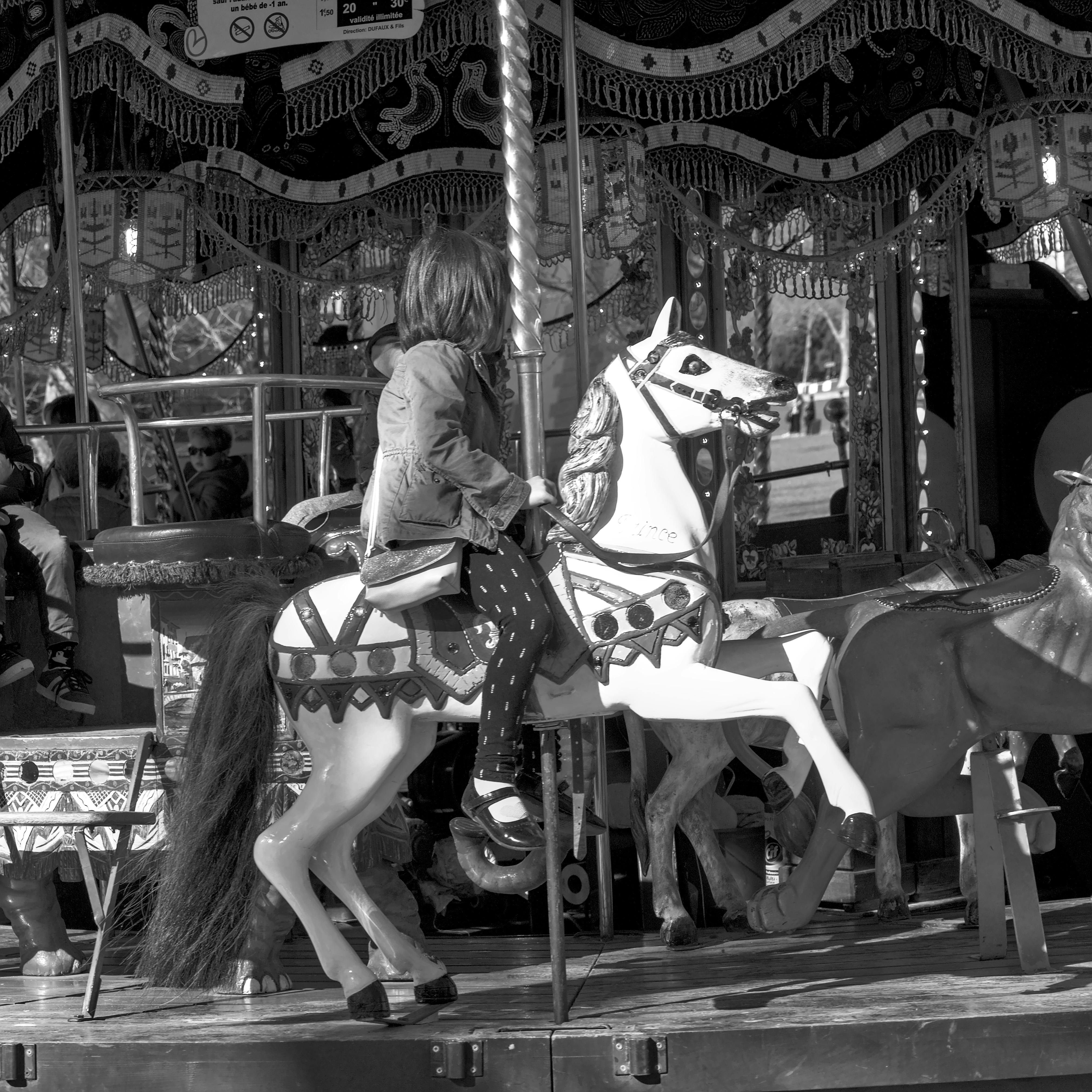 Bezpłatne Małe dziecko jedzie na zabytkowym koniku karuzeli w tętniącej życiem scenerii plenerowej, uchwycone w monochromatycznej kolorystyce. Zdjęcie z galerii